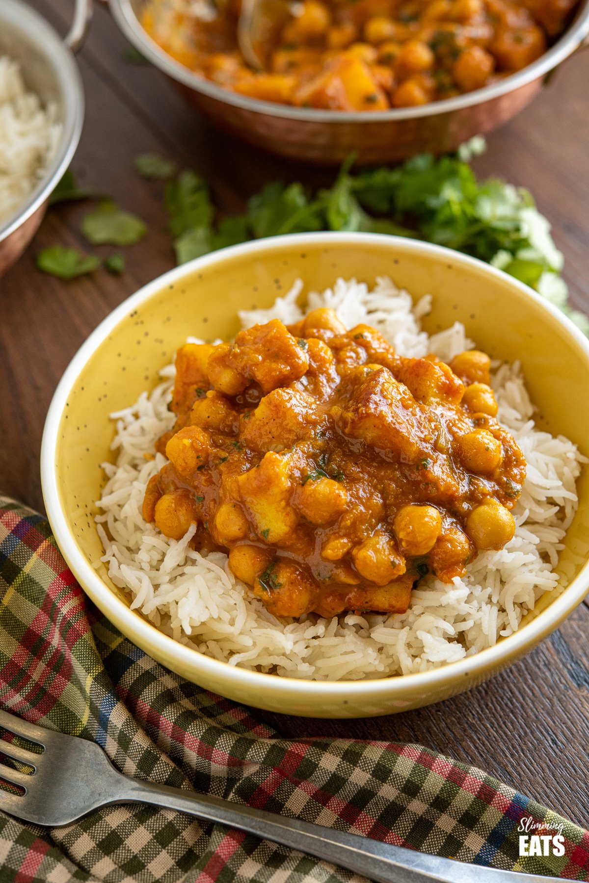 Chana Paneer over white basmati rice in yellow bowl with dishes in background with scattered cilantro and fork in front. 