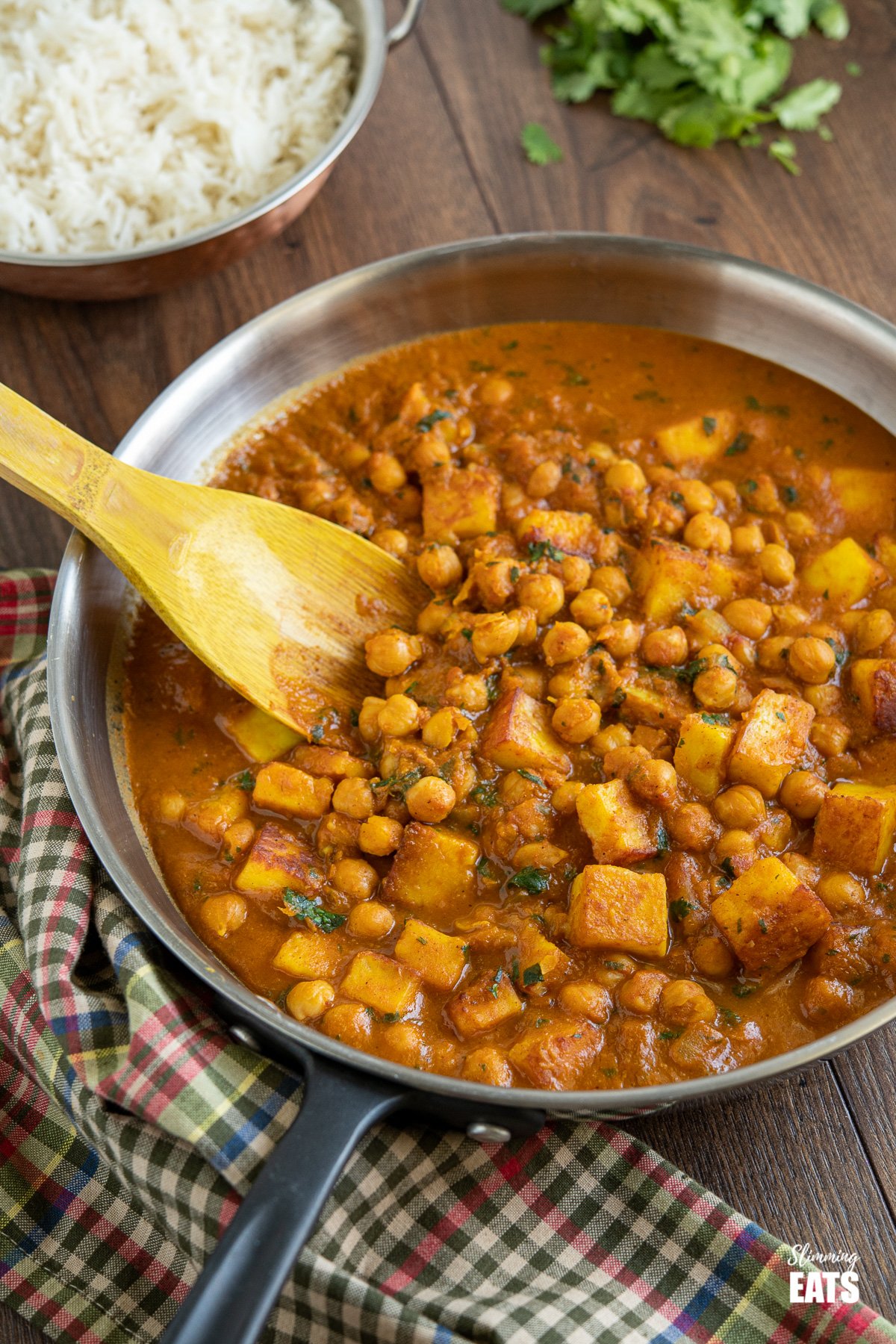 Chana Paneer in black handled frying pan with wooden spoon, rice and cilantro in the background