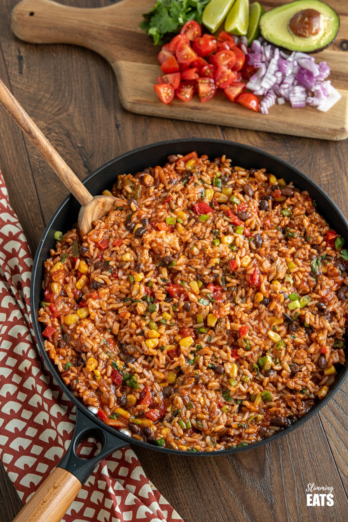 turkey burrito rice in black frying pan with wooden spoon, chopping board with toppings in background