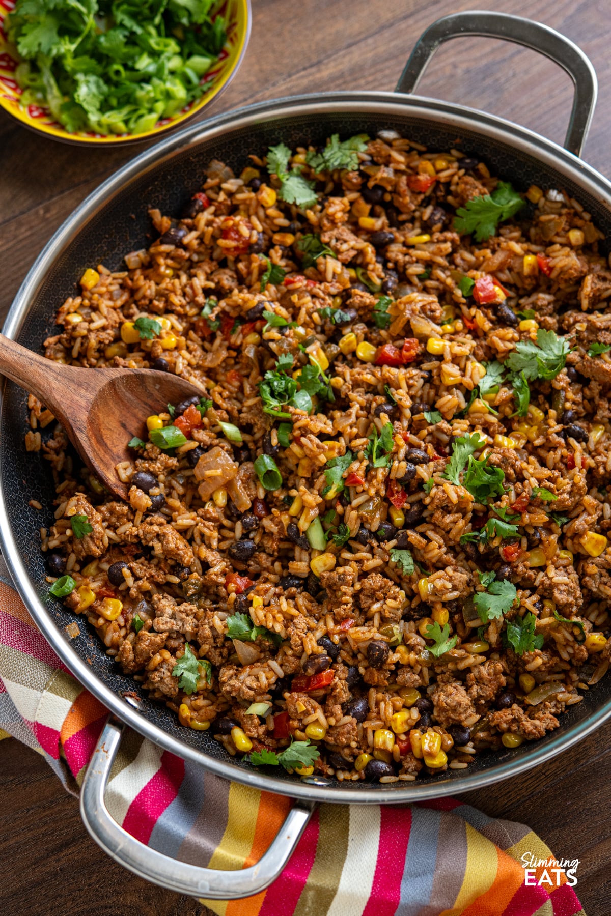 These Turkey Rice Burrito Bowls make a great family meal - sit down and grab a bowl with your favourite toppings. Gluten free, dairy free, Slimming Eats and Weight Watchers friendly One Pan Turkey Burrito Rice in a double handled non stick skillet next to a yellow and red bowl with chopped cilantro and green onions