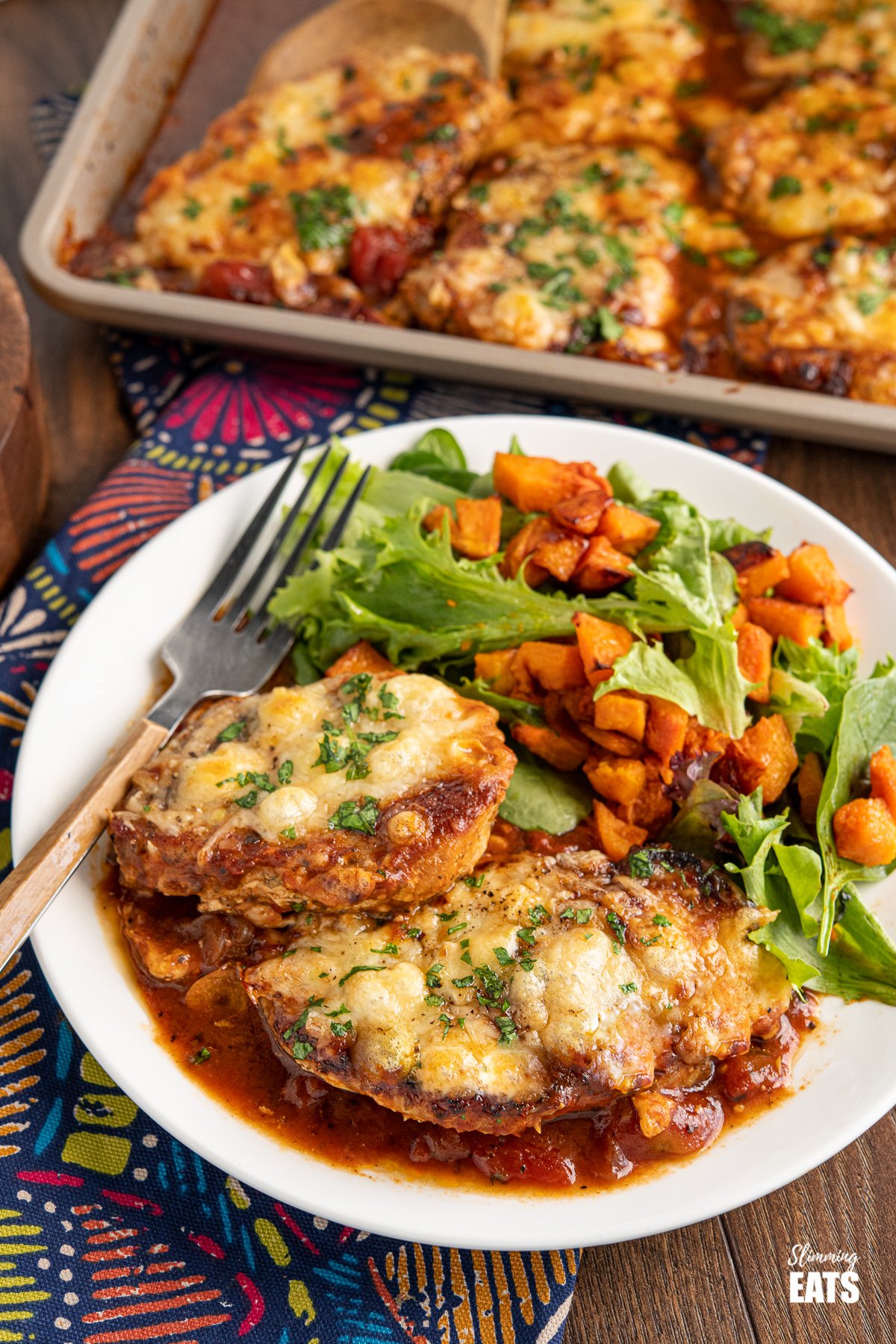 Cheesy Topped Mediterranean Pork Loin Chops - tender pork loin chops in a rich mediterranean style sauce topped with cheese and broiled until melted and golden. Gluten Free, Slimming Eats and Weight Watchers friendly white plate of Cheesy Topped Mediterranean Pork Loin Chops and roasted butternut squash with baby green, fork to the left of plate.