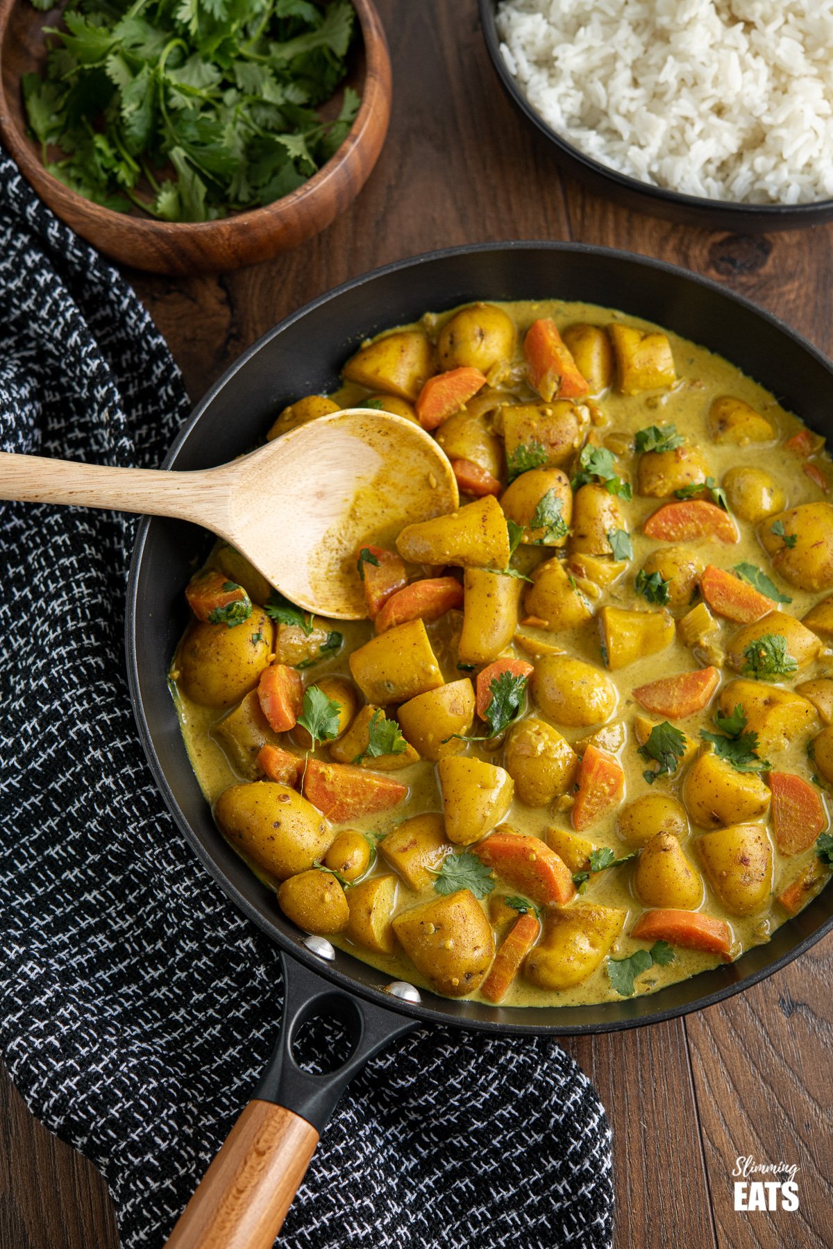 Creamy Coconut Potato Curry in a black frying pan with wooden spoon, bowls or rice and coriander in background