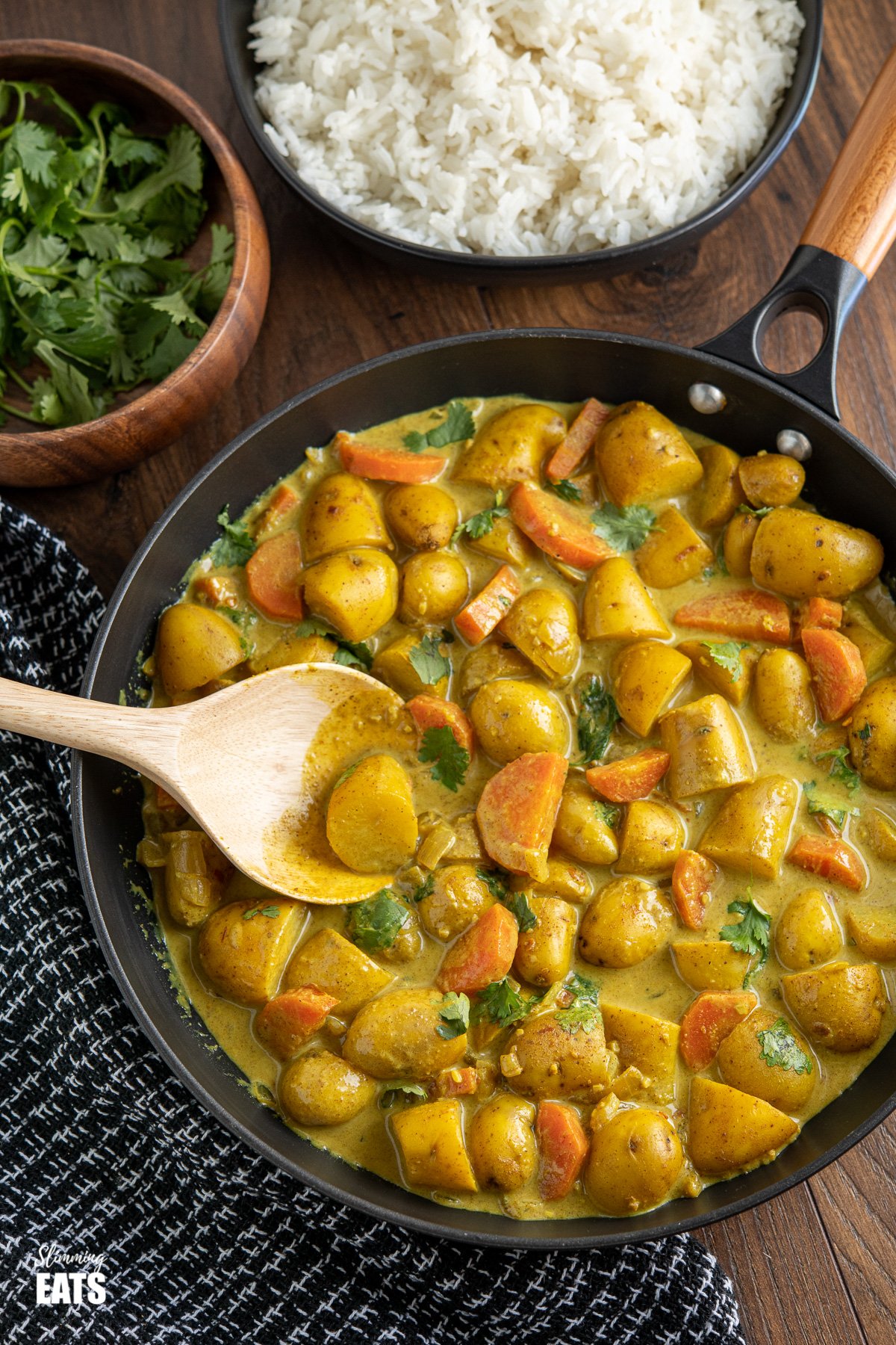 black frying pan with wooden handle filled with creamy coconut potato curry, bowls of rice and coriander in the background 