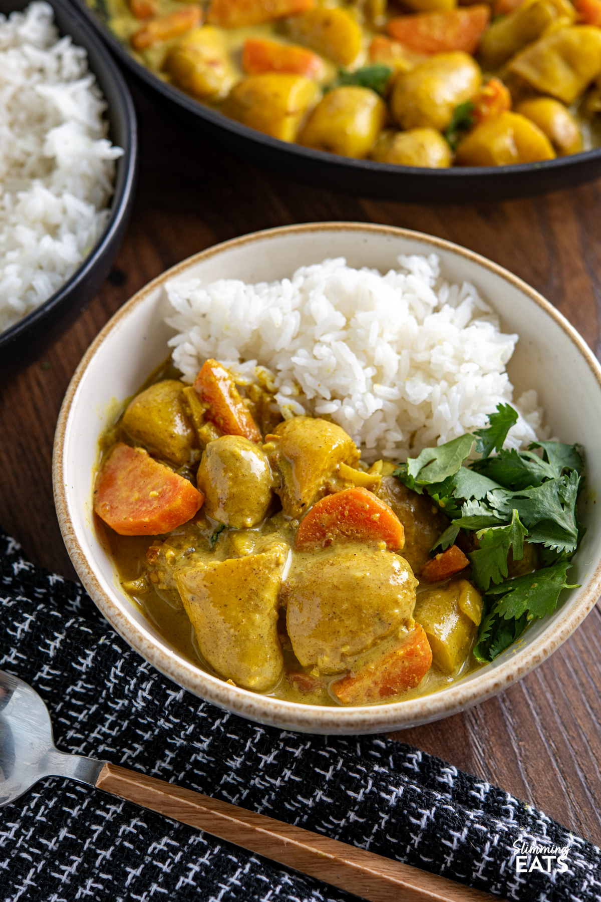 A simple and comforting Creamy Coconut Potato Curry with tender baby potatoes, carrots and warm spices in a rich creamy coconut sauce. serving of Creamy Coconut Potato Curry in a white bowl served wit rice with beige rim, spoon placed on black and white placement in front