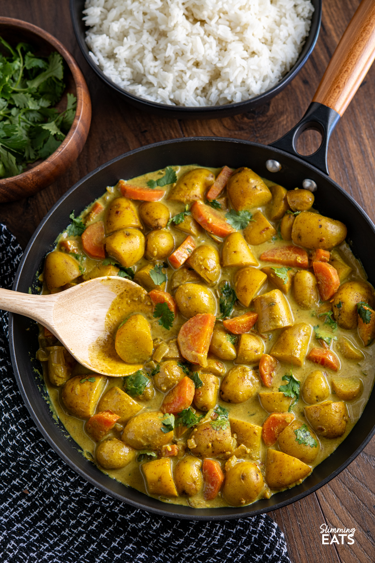 A simple and comforting Creamy Coconut Potato Curry with tender baby potatoes, carrots and warm spices in a rich creamy coconut sauce. Creamy Coconut Potato Curry in a black non stick skillet with wooden handle, bowl of coriander and rice visible