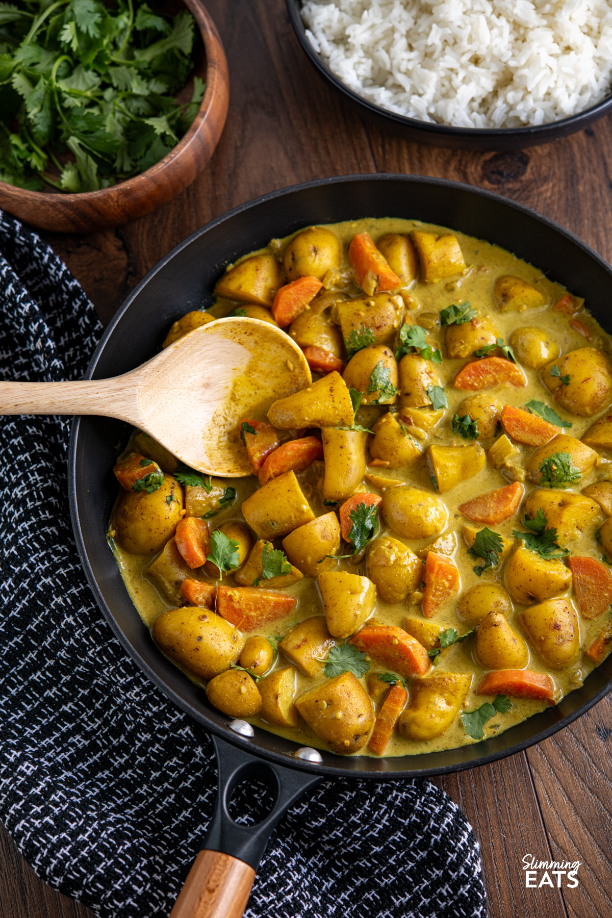 A simple and comforting Creamy Coconut Potato Curry with tender baby potatoes, carrots and warm spices in a rich creamy coconut sauce. Creamy Coconut Potato Curry in a black non stick skillet with wooden handle, bowl of coriander and rice visible