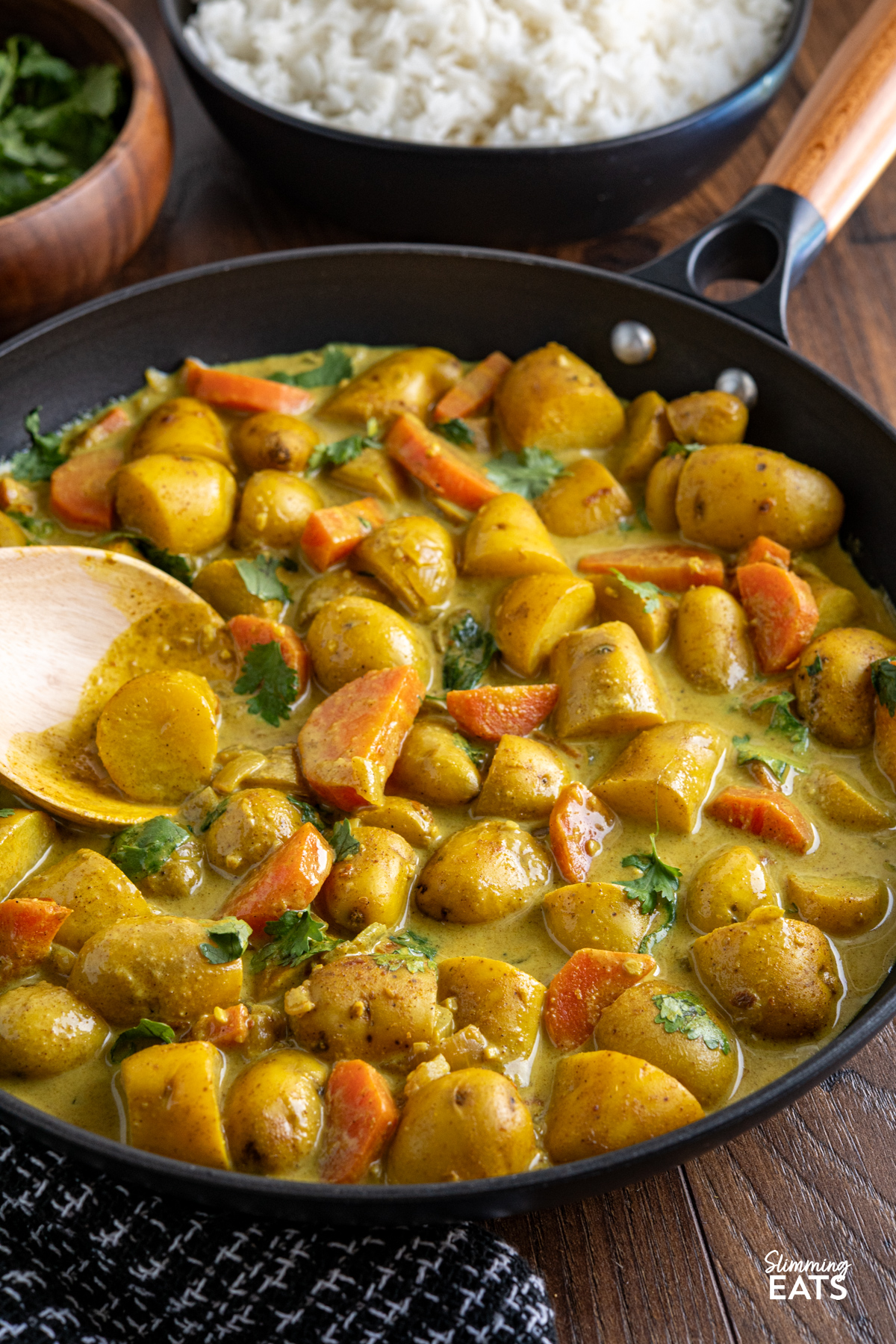 A simple and comforting Creamy Coconut Potato Curry with tender baby potatoes, carrots and warm spices in a rich creamy coconut sauce. Creamy Coconut Potato Curry in a black non stick skillet with wooden handle, bowl of coriander and rice visible