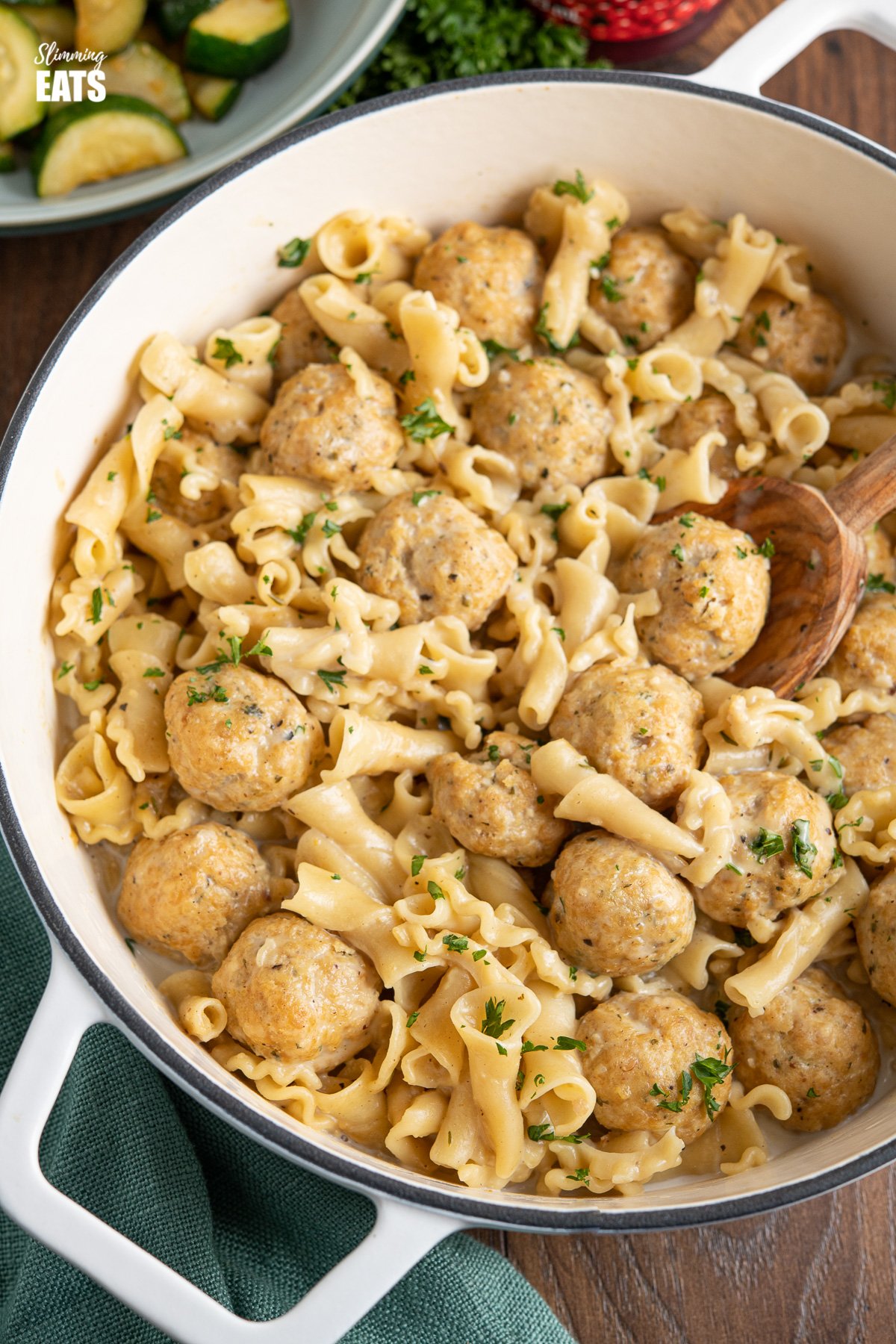 One Pot Turkey Swedish Meatball Pasta in white cast iron pan with olive wood spoon with bowl of zucchini in background