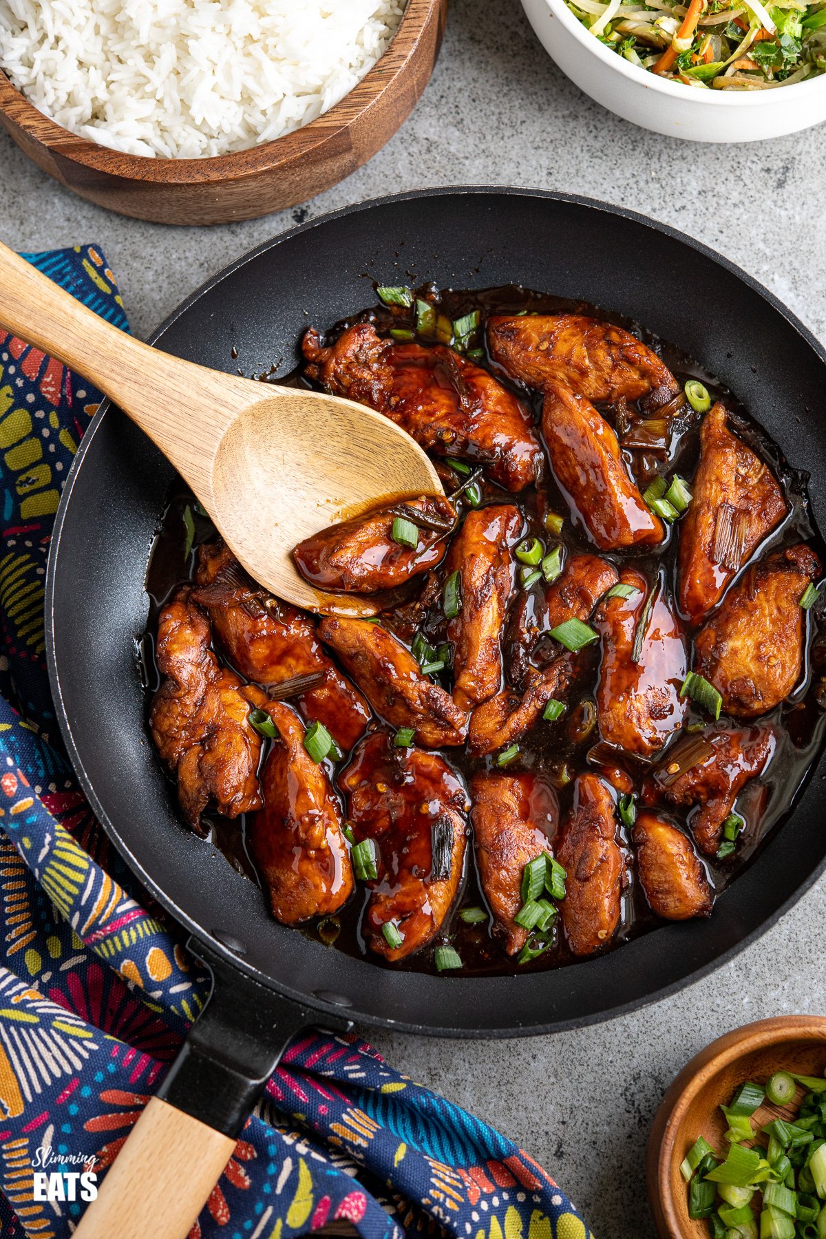 asian sprite chicken in a wooden handled black frying pan with bowl of rice and stir fried veg in background