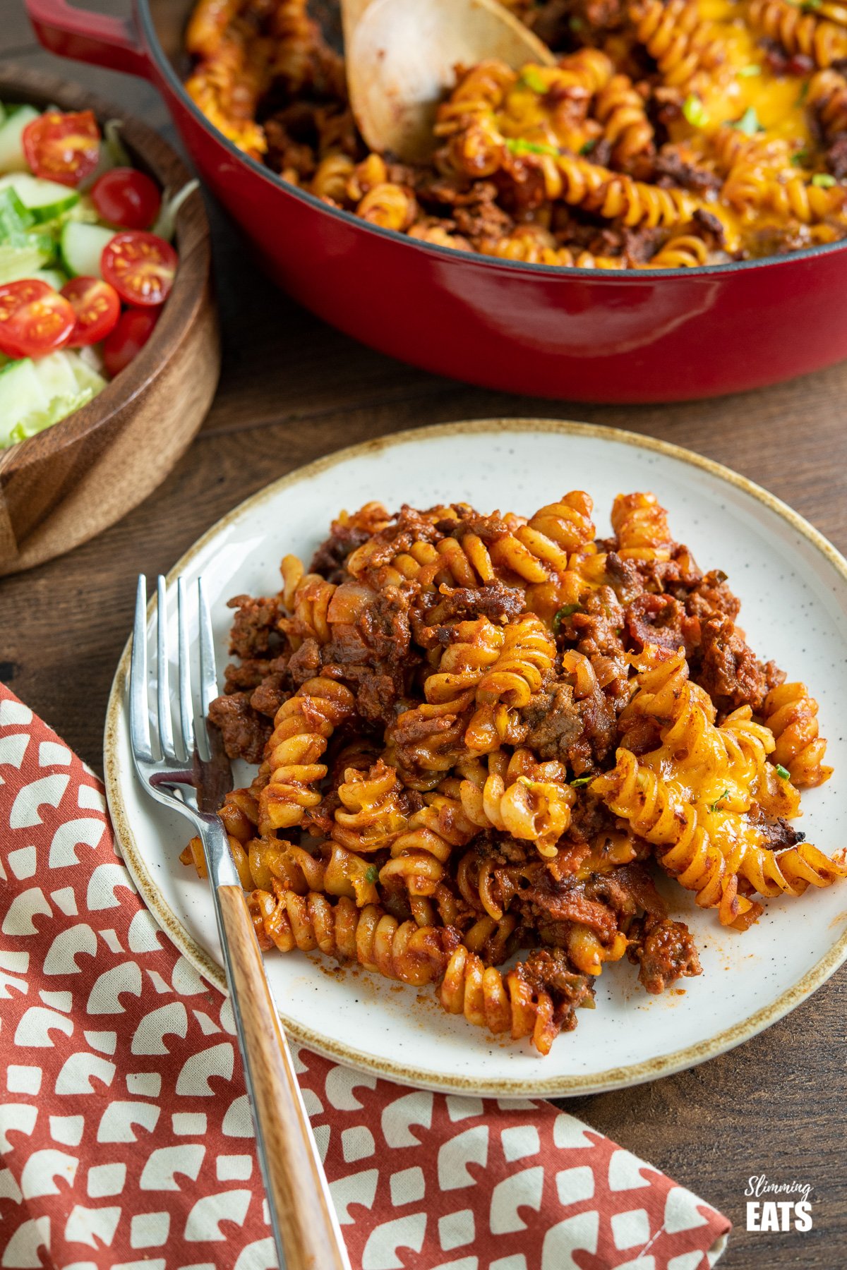 serving of BBQ Bacon Cheeseburger Pasta Bake on white plate with beige rim, fork placed to the left and a wooden bowl of salad and cast iron skillet filled with the pasta in the background