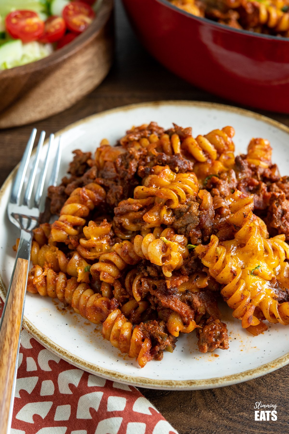 close up of serving of BBQ Bacon Cheeseburger Pasta Bake on white plate with beige rim, fork placed to the left and a wooden bowl of salad and red cast iron skillet filled in the background