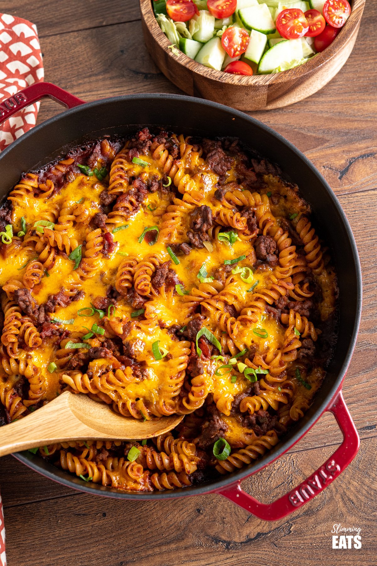 BBQ Bacon Cheeseburger Pasta Bake in red cast iron skillet with wooden spoon and wooden bowl of mixed salad in the background