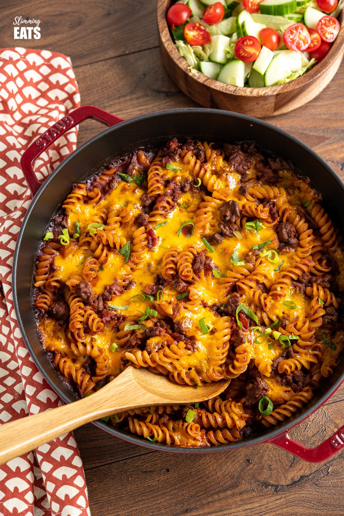 BBQ Bacon Cheeseburger Pasta Bake in red cast iron skillet with wooden spoon, and wooden bowl of salad in background