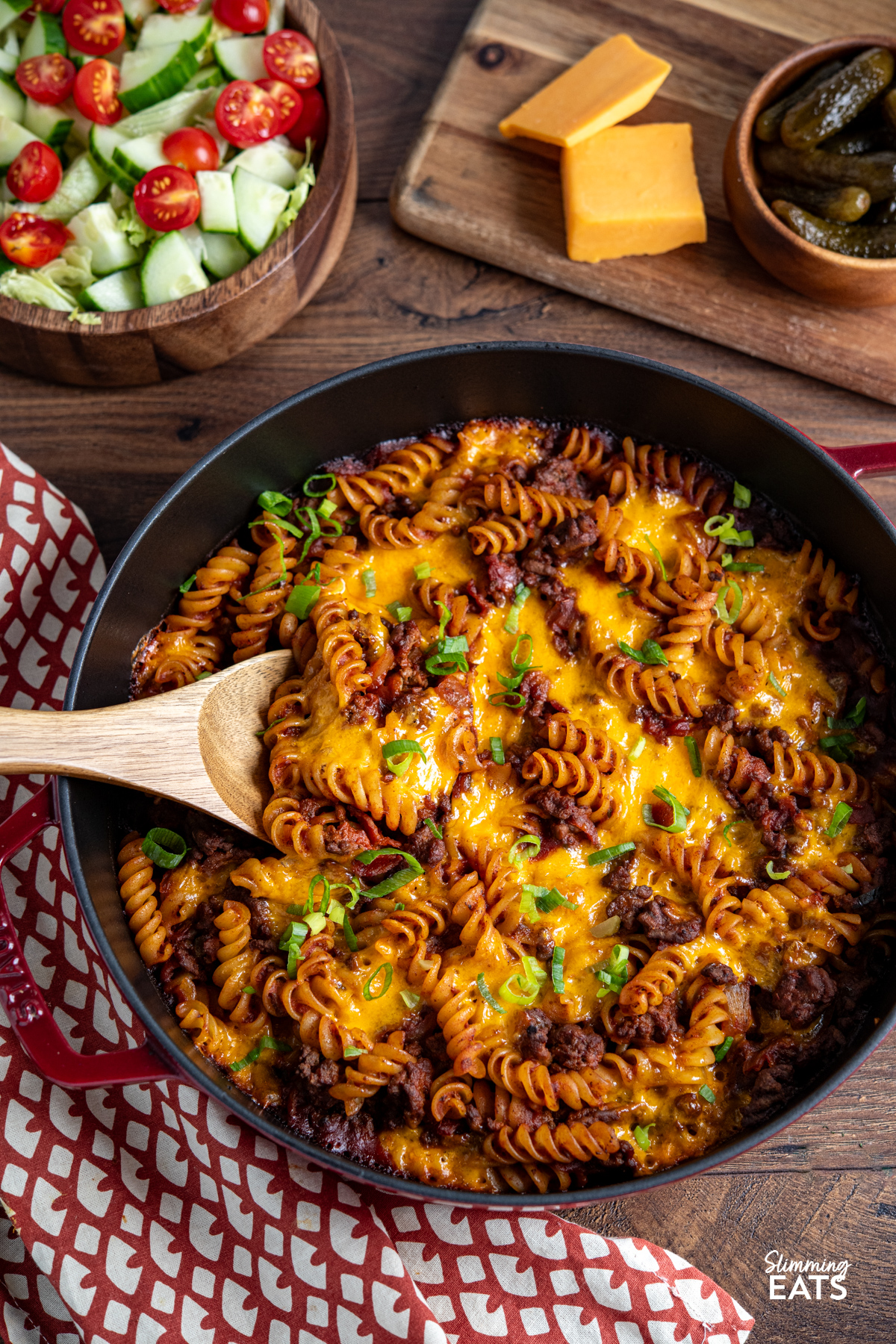 This BBQ Bacon Cheeseburger Pasta Bake is a hearty, flavour-packed family dinner with all the best cheeseburger flavours combined in one cheesy, comforting pasta dish. BBQ Bacon Cheeseburger Pasta Bake in cherry cast iron staub skillet, with wooden bowl of salad in background and board with cheddar and pickles