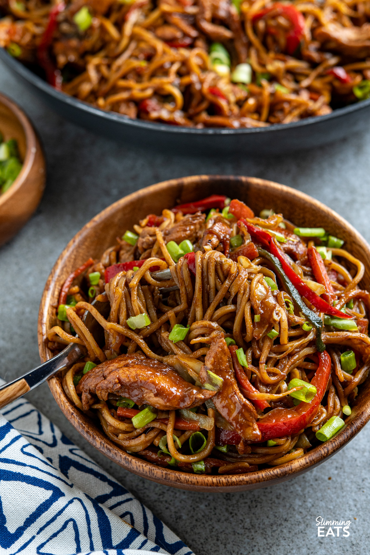 serving of hoisin chicken noodles in wooden bowl with fork place to left in bowl and frying pan of noodles in background