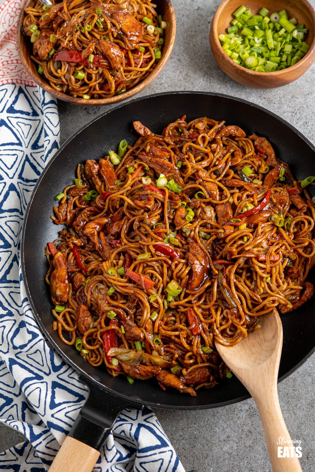 Hoisin chicken noodles in black frying pan with wooden handle, bowl of noodles and spring onions in background