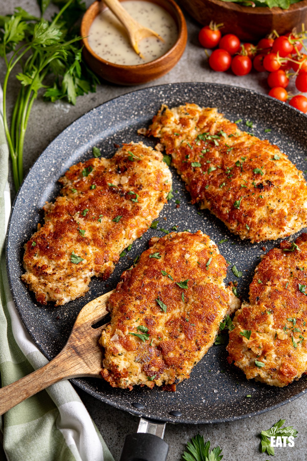 Four Golden Parmesan Crusted Chicken on frying pan with wooden spatula, tomatoes, dressing and salad in background