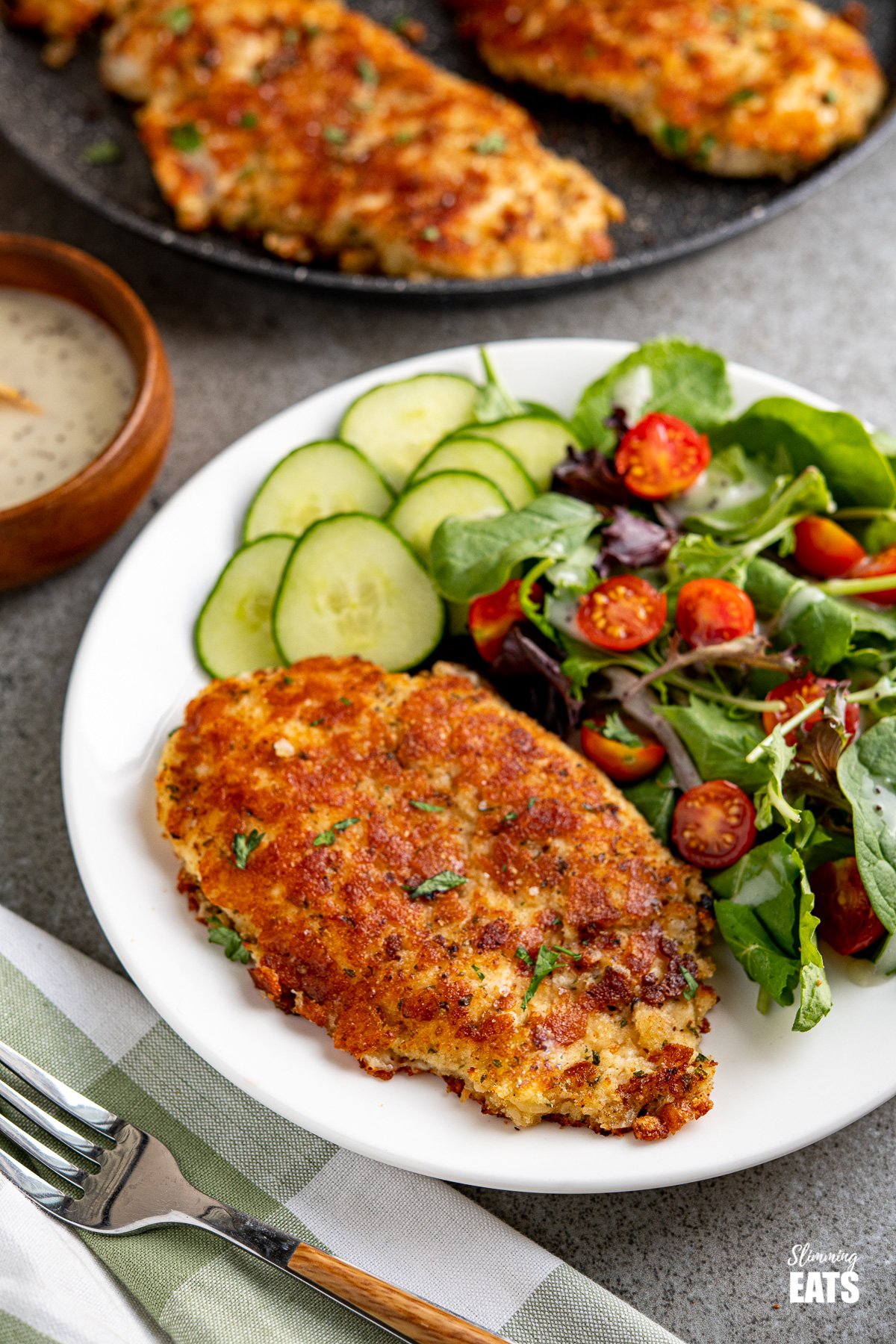 Golden Parmesan Crusted Chicken cutlet on white plate with baby greens, tomatoes and cucumber, fork placed to the side and skillet bowl of dressing background