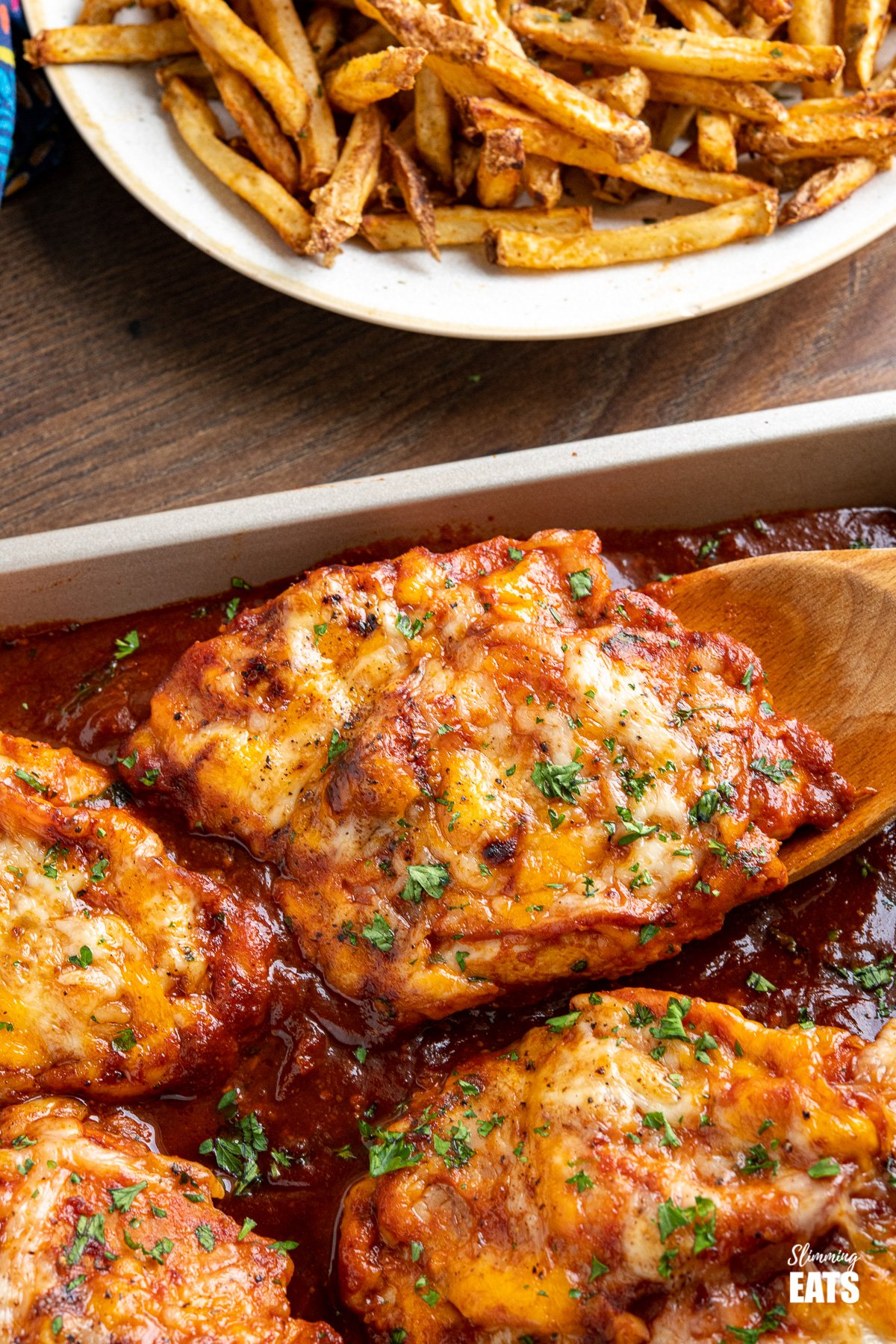 Hunters Chicken - tender chicken breasts in a delicious homemade barbecue sauce with slices of smoky bacon, all topped with cheese and baked until melted and golden. #chicken #barbecue #cheese #bacon #slimmingeats #weightwatchers #smartpoints Hunters Chicken on baking tray with wooden spatula with oven baked fries on plate in background