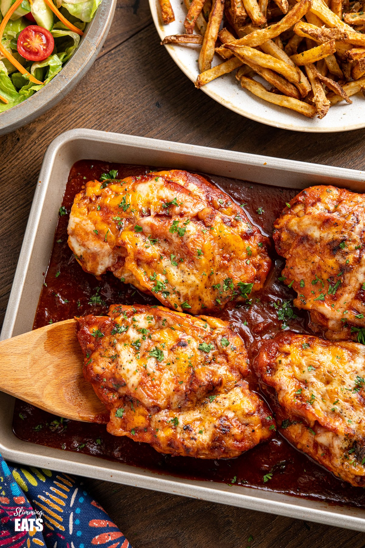 Hunters Chicken on baking tray with wooden spatula, oven baked fries and bowl of salad in background