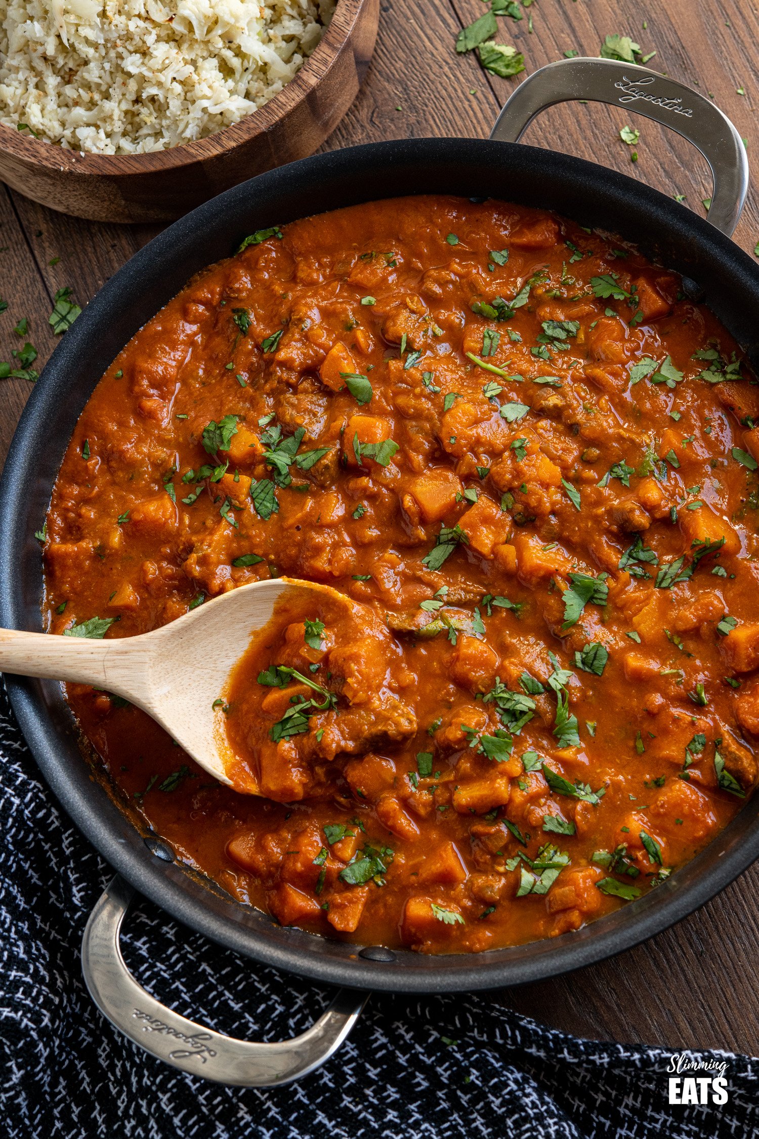 Beef and Sweet Potato Tikka Masala in a black pan with wooden spoon and wooden bowl with cauliflower rice with scattered coriander