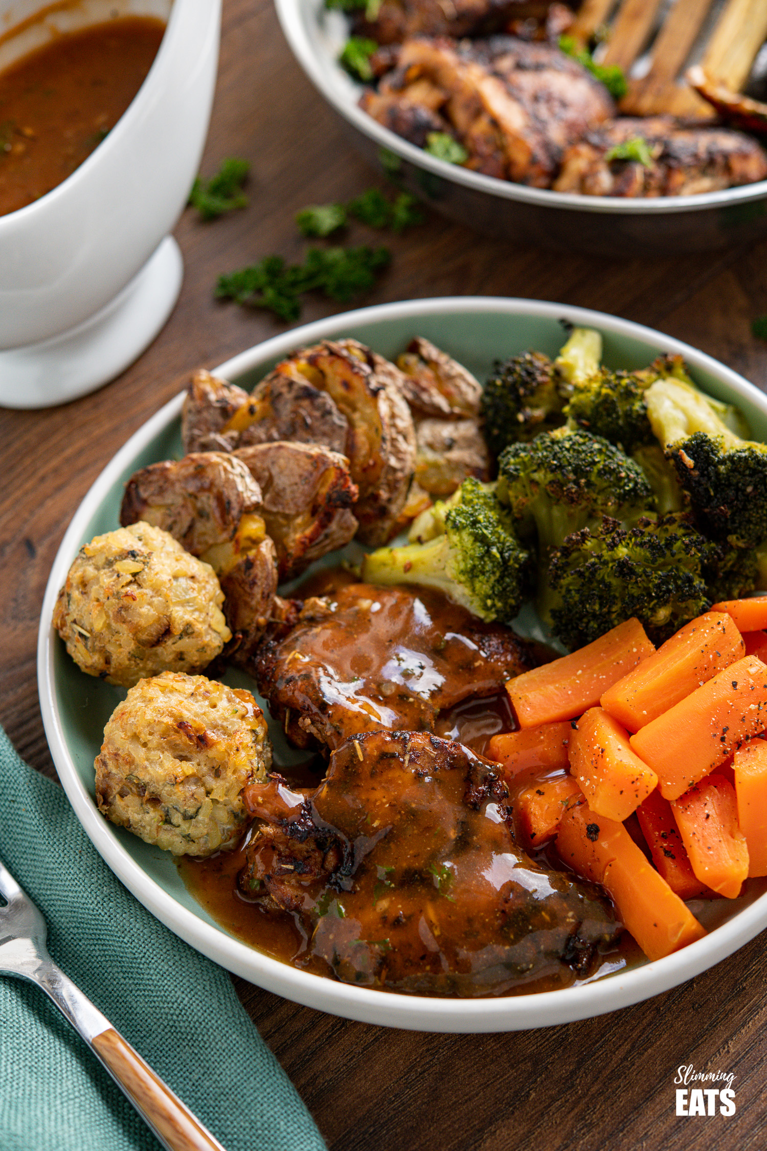 Seasoned Chicken with Rich Gravy on teal plate with stuffing balls, crushed potatoes, broccoli and carrots, jug of gravy in background.