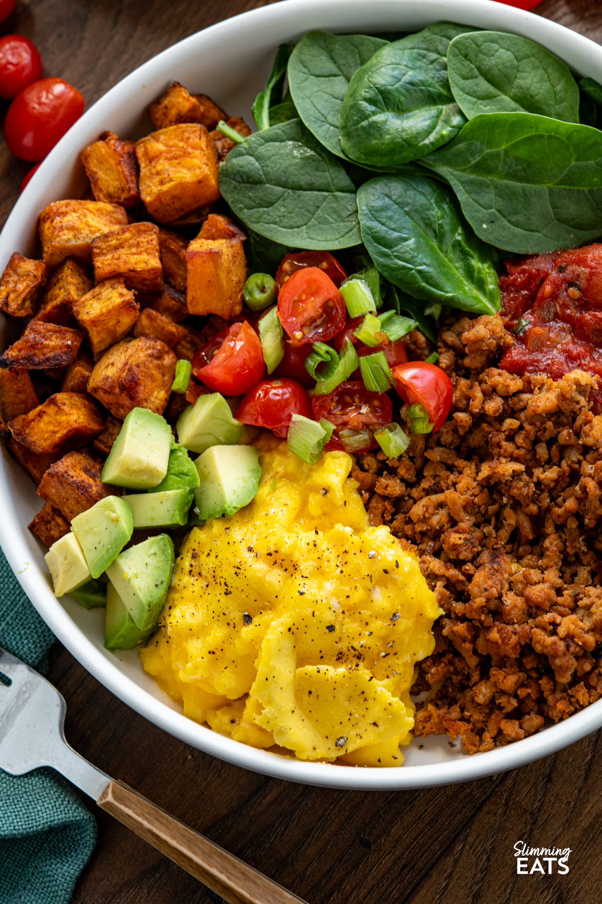Taco Breakfast Bowl - lean seasoned ground turkey with sweet potato, spicy tomatoes, spinach, avocado and perfectly scrambled eggs for a filling high protein start to the day. #slimmingeats #weightwatchers #taco #breakfast #eggs #glutenfree #dairyfree #paleo #whole30 close up of turkey taco meat, eggs, spinach, sweet potato, avocado, tomatoes in a bowl with additional bowl in background with scattered tomatoes and half an avocado