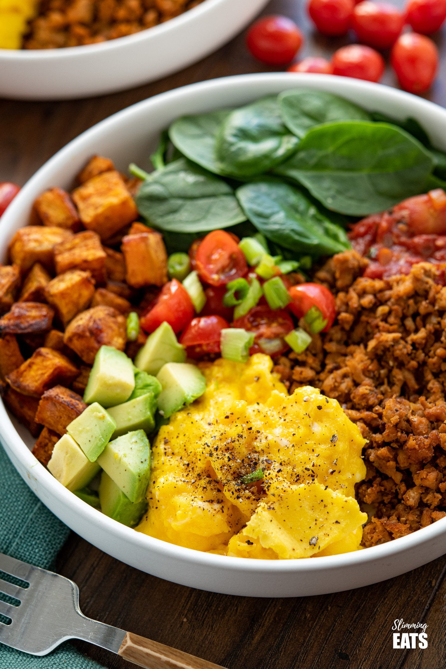 turkey taco meat, eggs, spinach, sweet potato, avocado, tomatoes in a bowl with additional bowl in background with scattered tomatoes and half an avocado