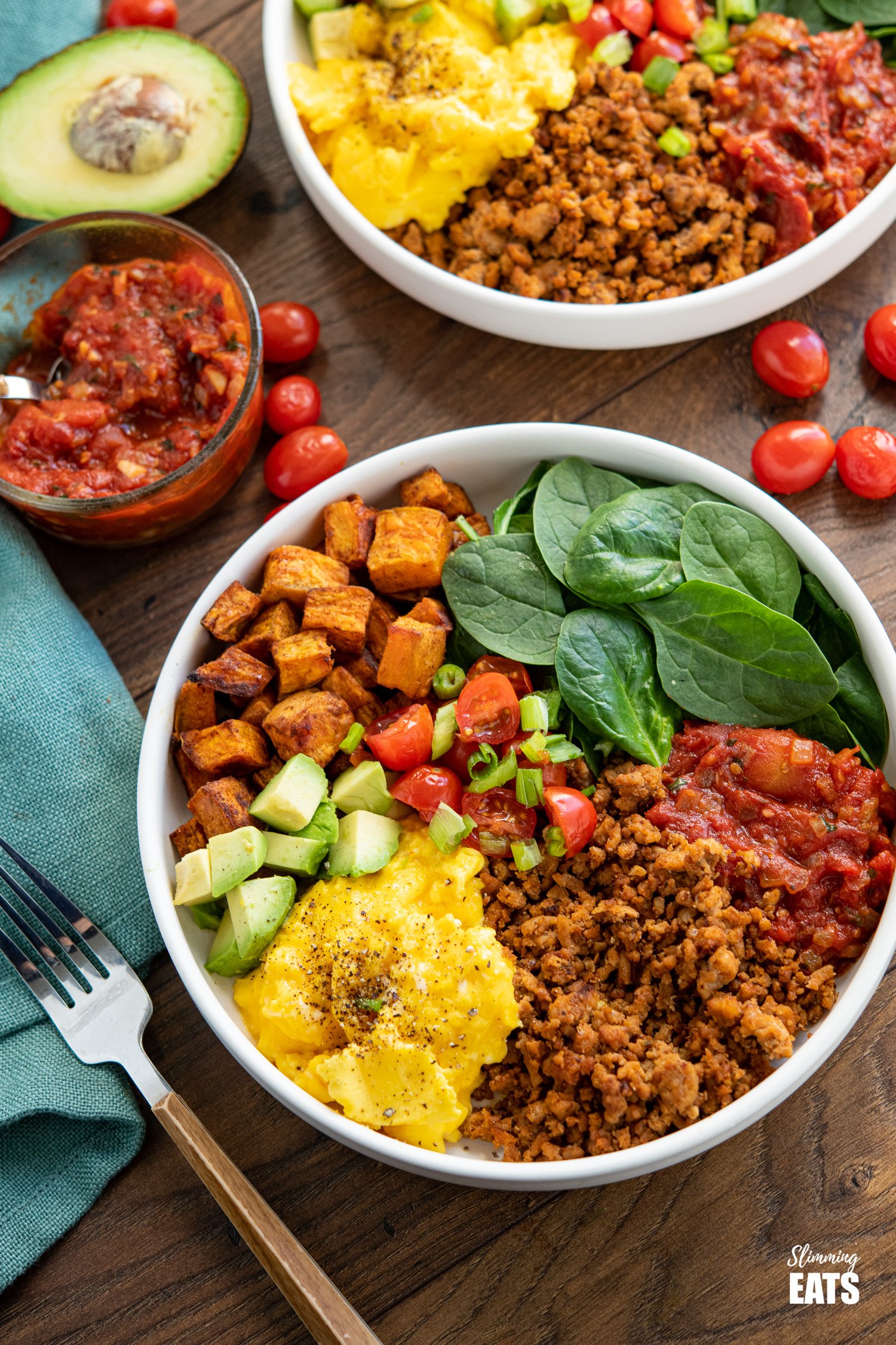 turkey taco meat, eggs, spinach, sweet potato, avocado, tomatoes in a bowl with additional bowl in background with scattered tomatoes and half an avocado