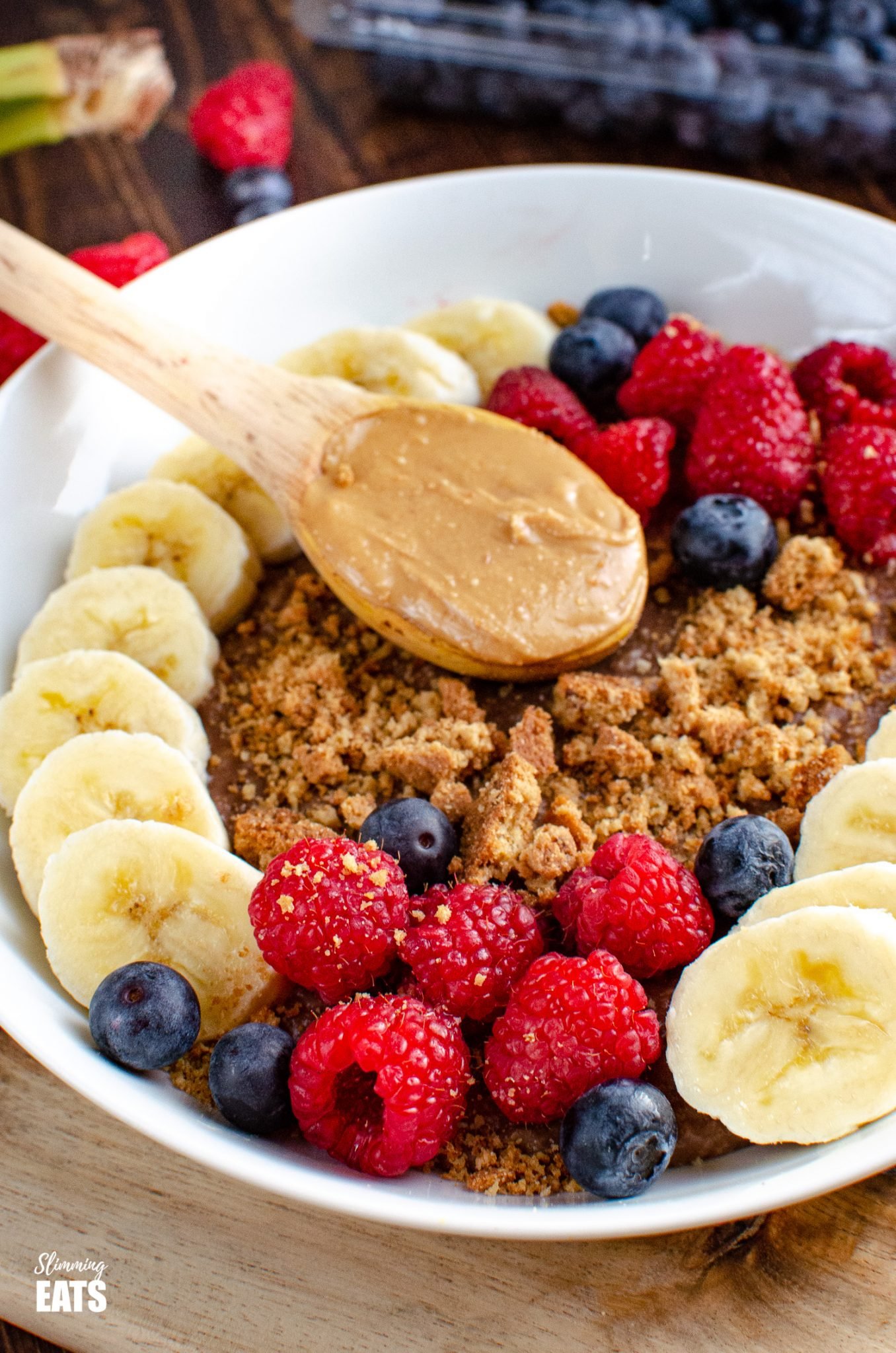 Chocolate Peanut Butter Oatmeal in white bowl with wooden spoon of peanut butter, slices of banana, raspberries, blueberries and a mini crumbled cookie