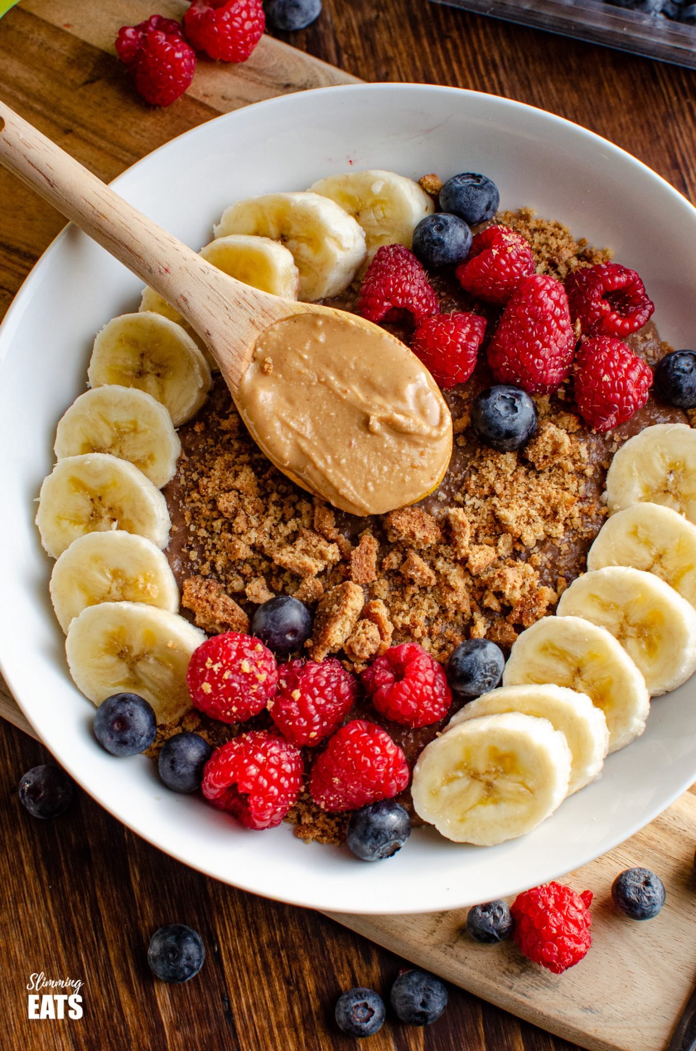 Chocolate Peanut Butter Oatmeal in white bowl with wooden spoon of peanut butter, slices of banana, raspberries, blueberries and a mini crumbled cookie