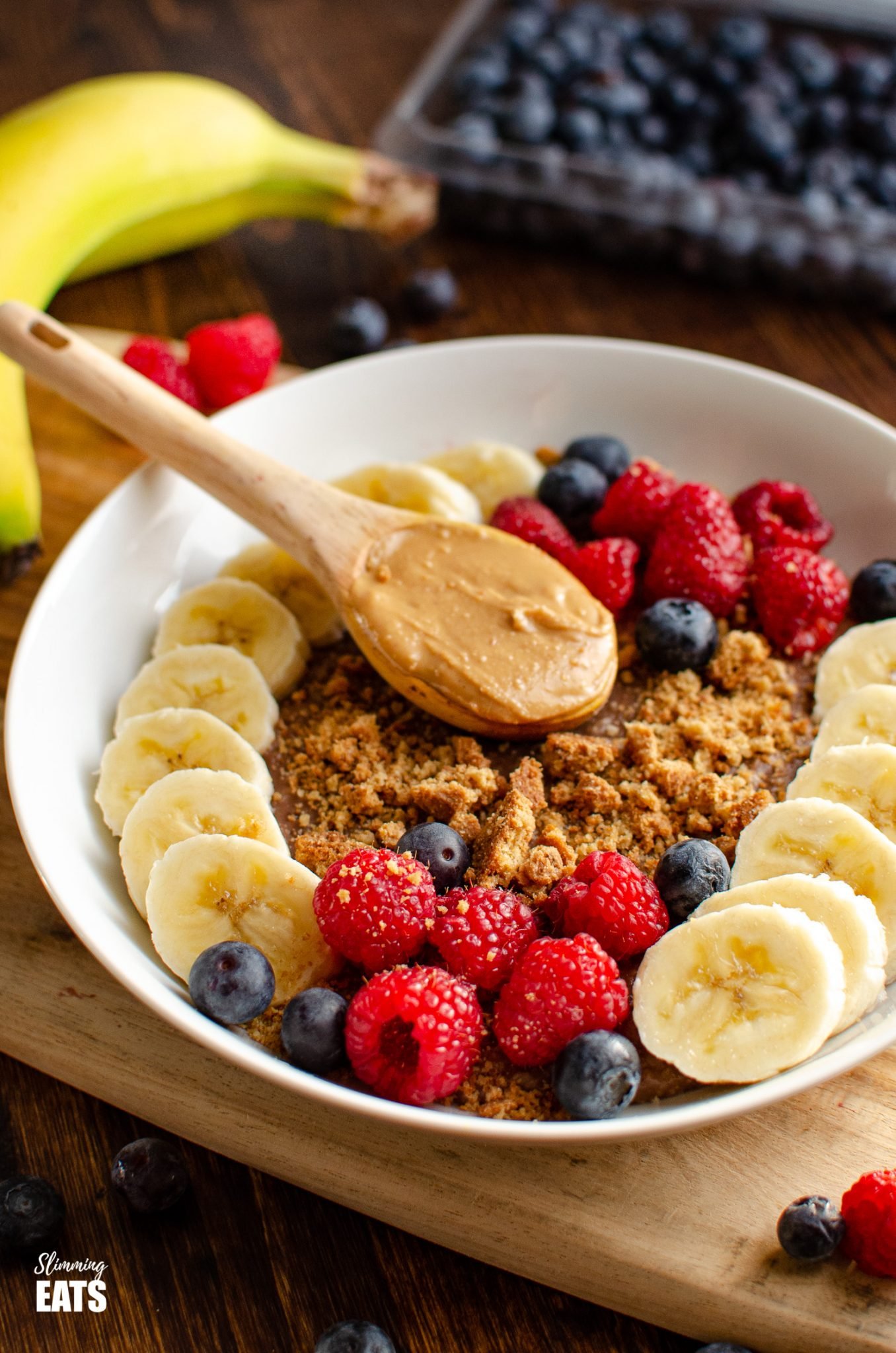 Chocolate Peanut Butter Oatmeal in white bowl with wooden spoon of peanut butter, slices of banana, raspberries, blueberries and a mini crumbled cookie