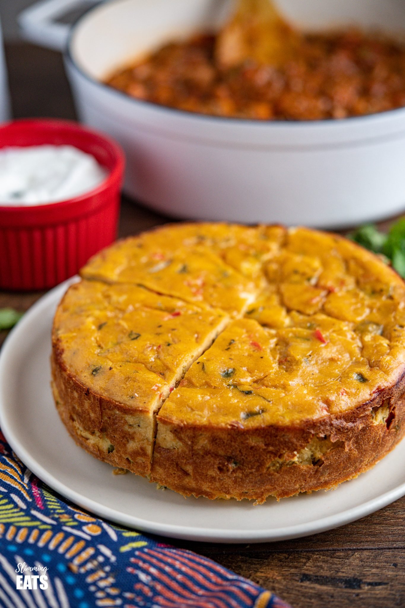 sliced lentil cheddar bake on white plate with cast iron pan of chilli in background with red dish of sour cream and scattered cilantro