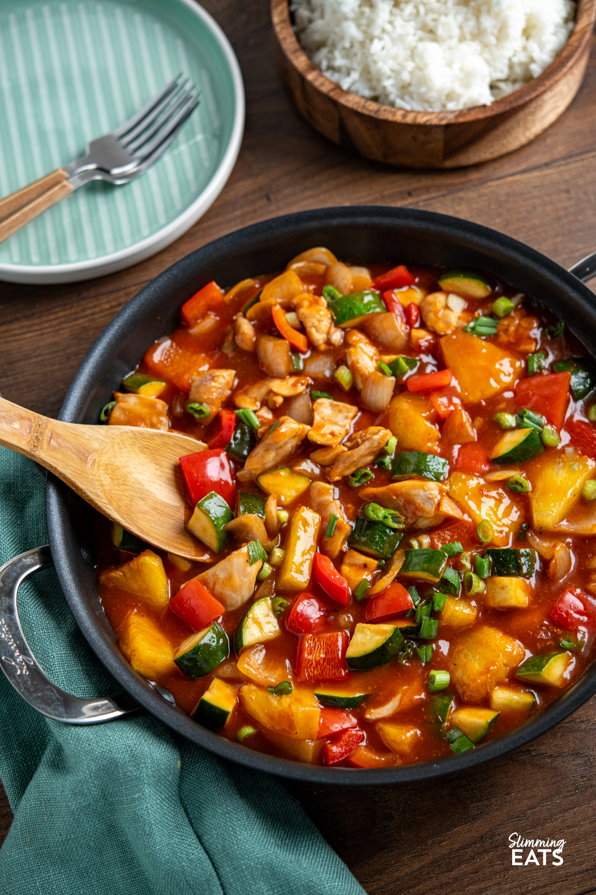 Sweet and Sour Chicken - a classic Chinese recipe you can make in your own home, with tender pieces of chicken thigh, fresh vegetables and pineapple in a delicious sauce. Dairy Free, Gluten Free, Slimming Eats and Weight Watchers friendly chinese sweet and sour chicken in metal handled pan with wooden bowl of rice and teal plate with forks in background