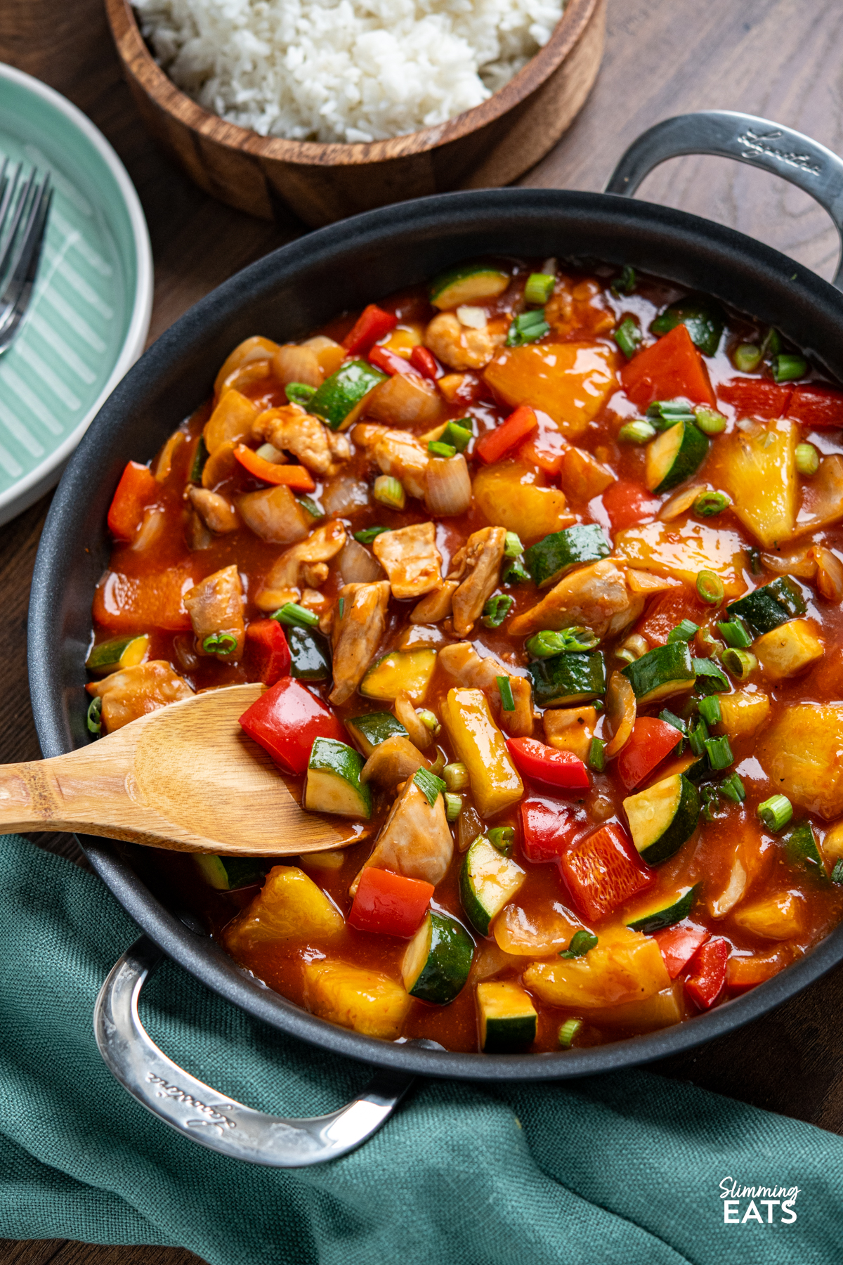 Sweet and Sour Chicken - a classic Chinese recipe you can make in your own home, with tender pieces of chicken thigh, fresh vegetables and pineapple in a delicious sauce. Dairy Free, Gluten Free, Slimming Eats and Weight Watchers friendly chinese sweet and sour chicken in metal handled pan with wooden bowl of rice in background