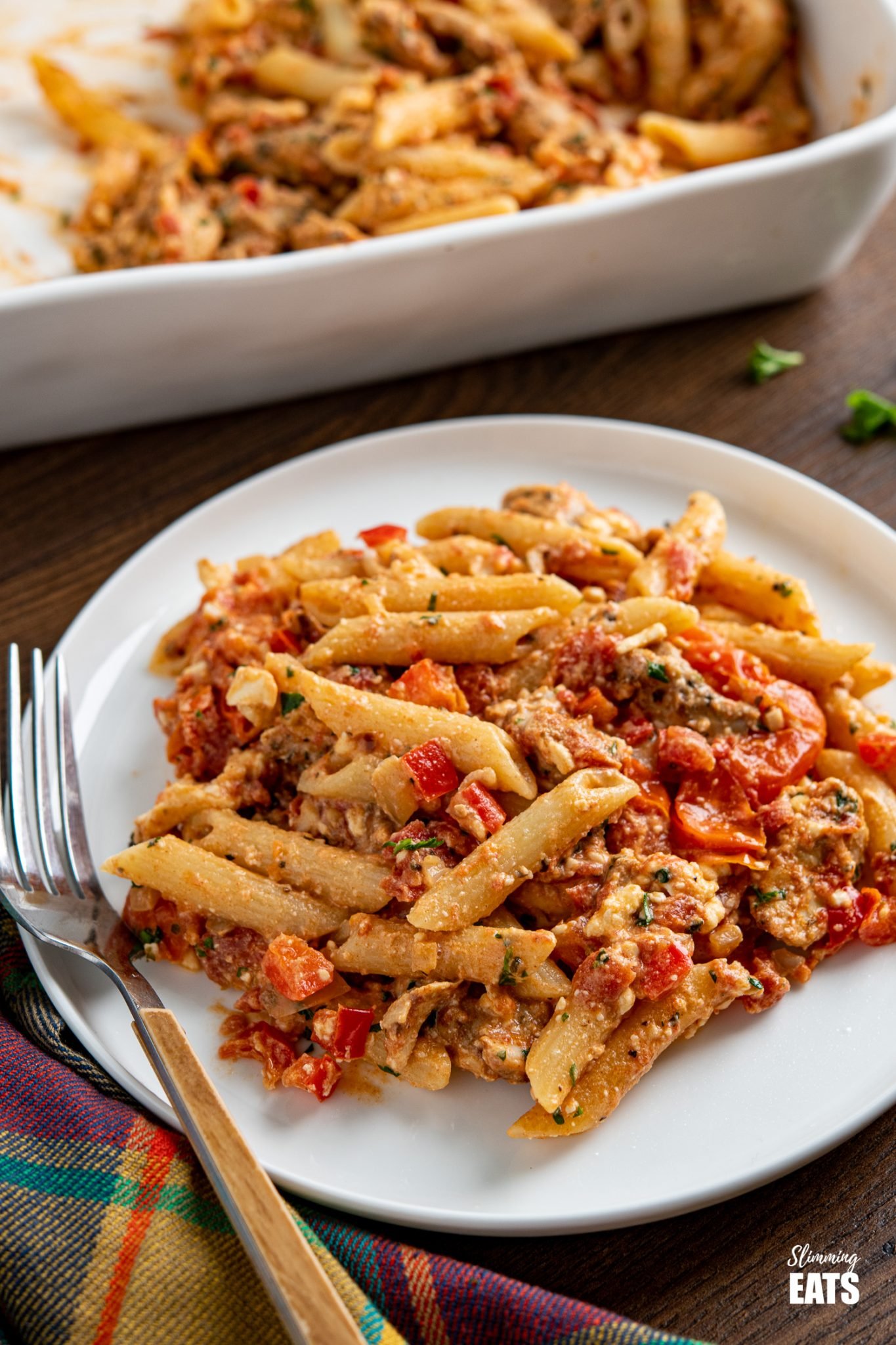 Baked feta pasta with chicken on white plate, baking dish in background and scattered herbs