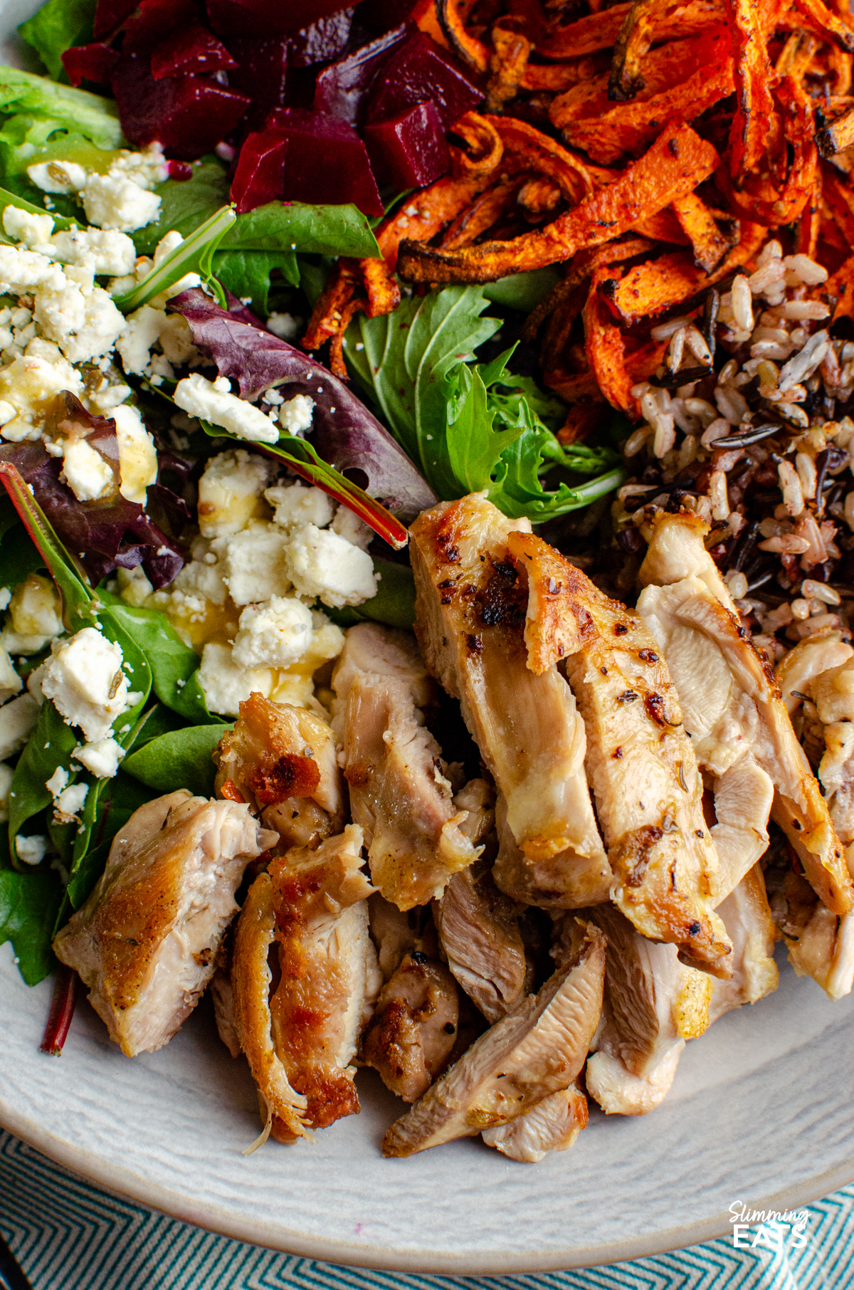 close up of Chicken Wild Rice Bowl - tender seasoned chicken thighs with a delicious nutty wild rice blend, roasted carrot strands, salad with beets and feta in a bowl 