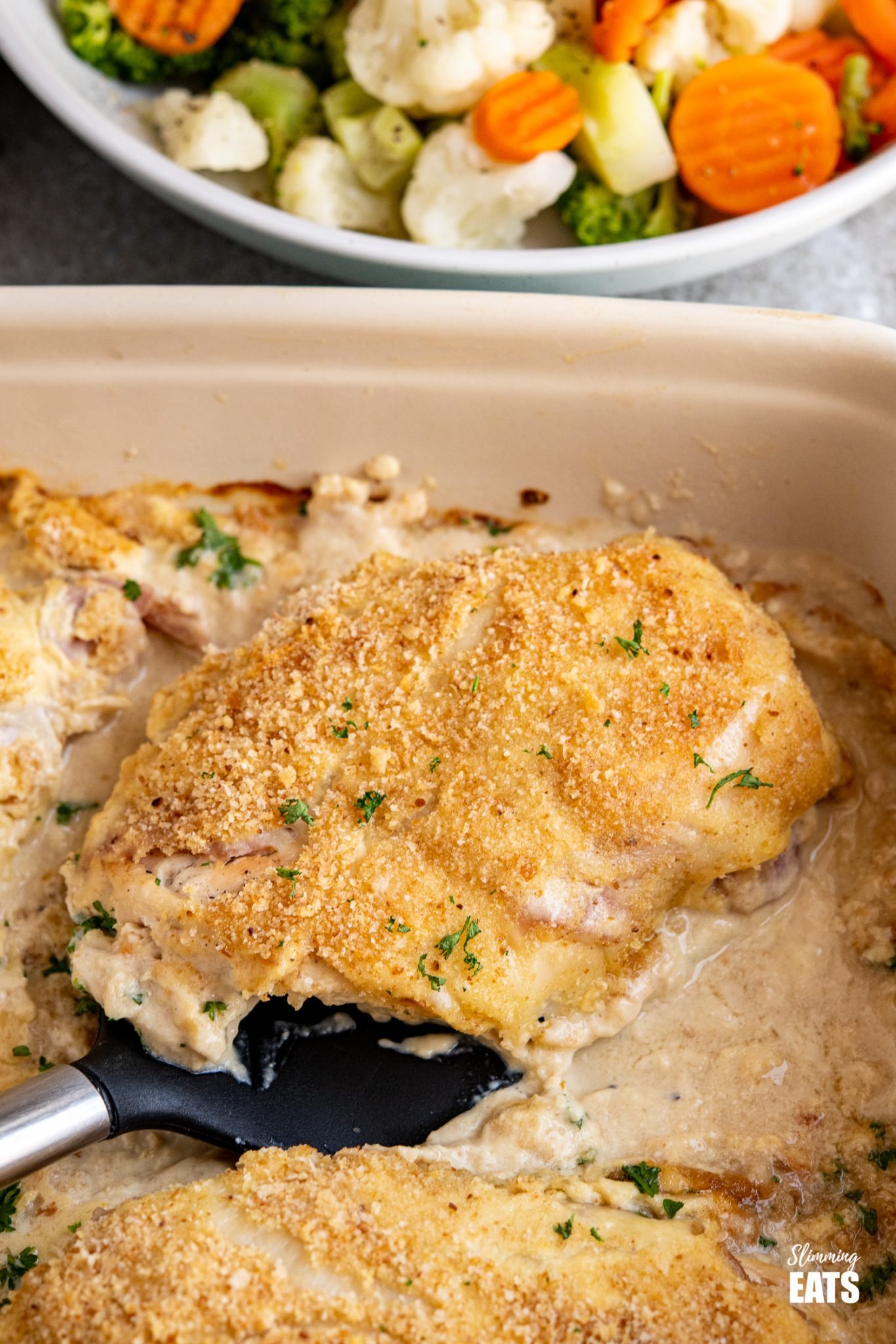 Easy Chicken Cordon Bleu - tender chicken breasts, topped with ham, swiss cheese, healthier dijon creamy sauce and breadcrumbs. Slimming World and Weight Watchers friendly close up of Easy Chicken Cordon Bleu on spatula in baking dish with bowl of steam veg above
