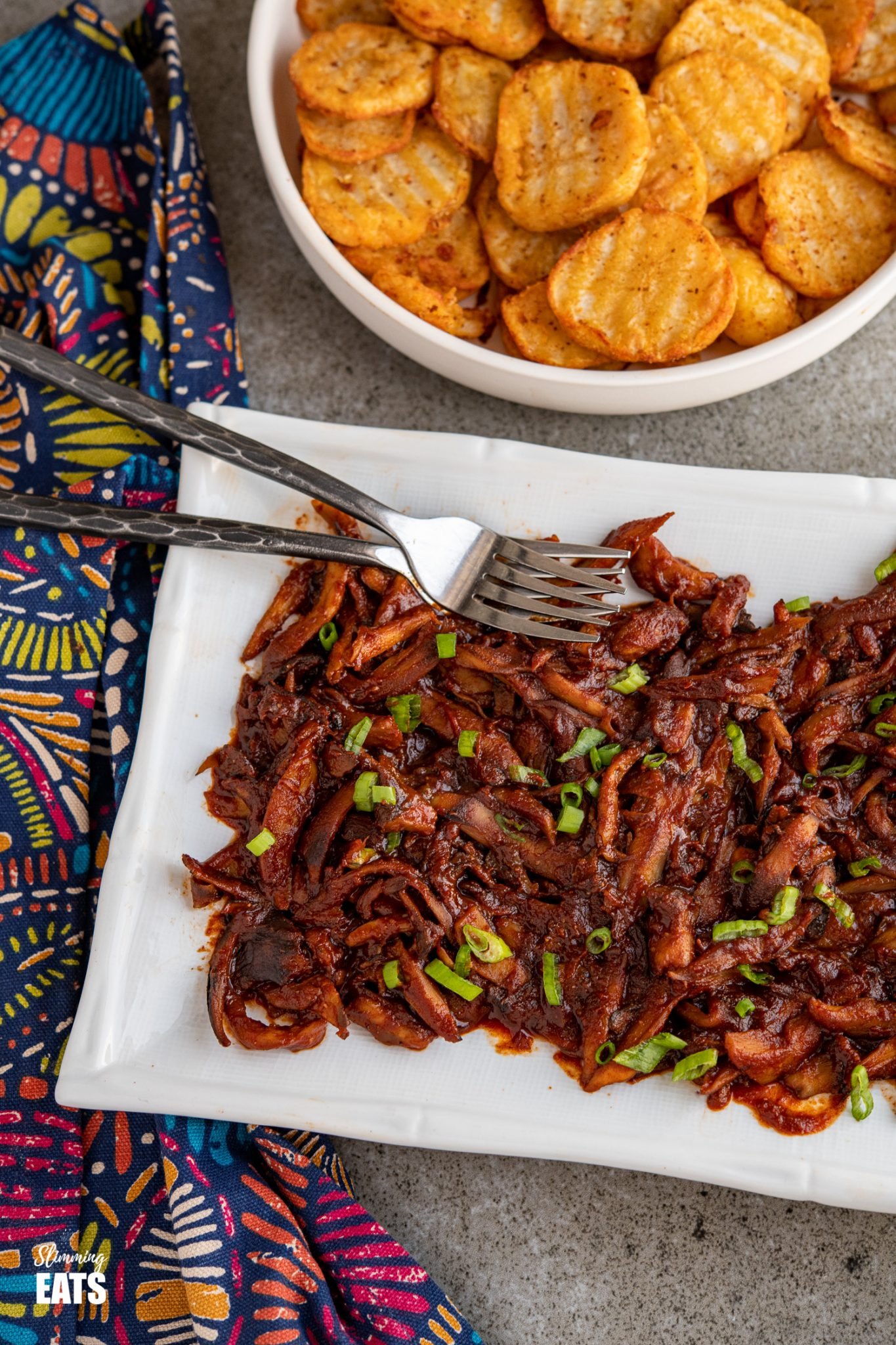 Pulled Mushrooms with BBQ Sauce on white rectangle plate with two forks with bowl of crispy potato slices in the background.