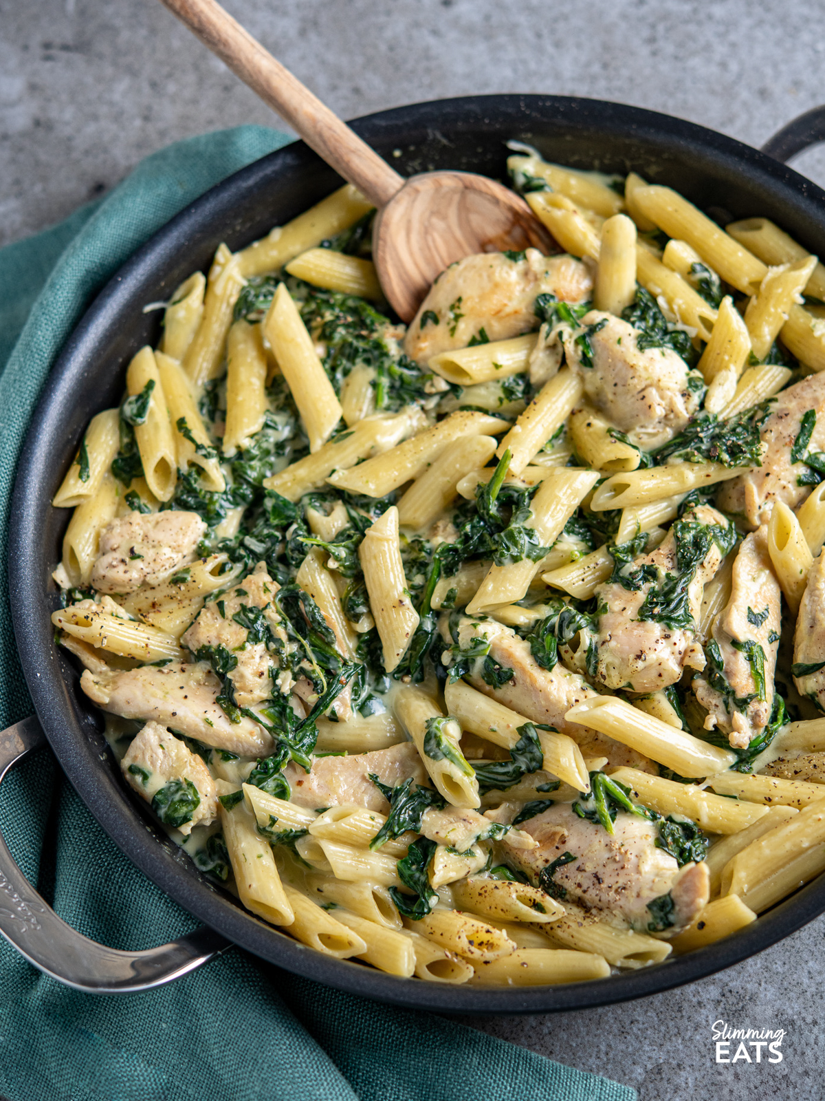 creamy garlic chicken with spinach penne pasta in black frying pan with wooden spoon