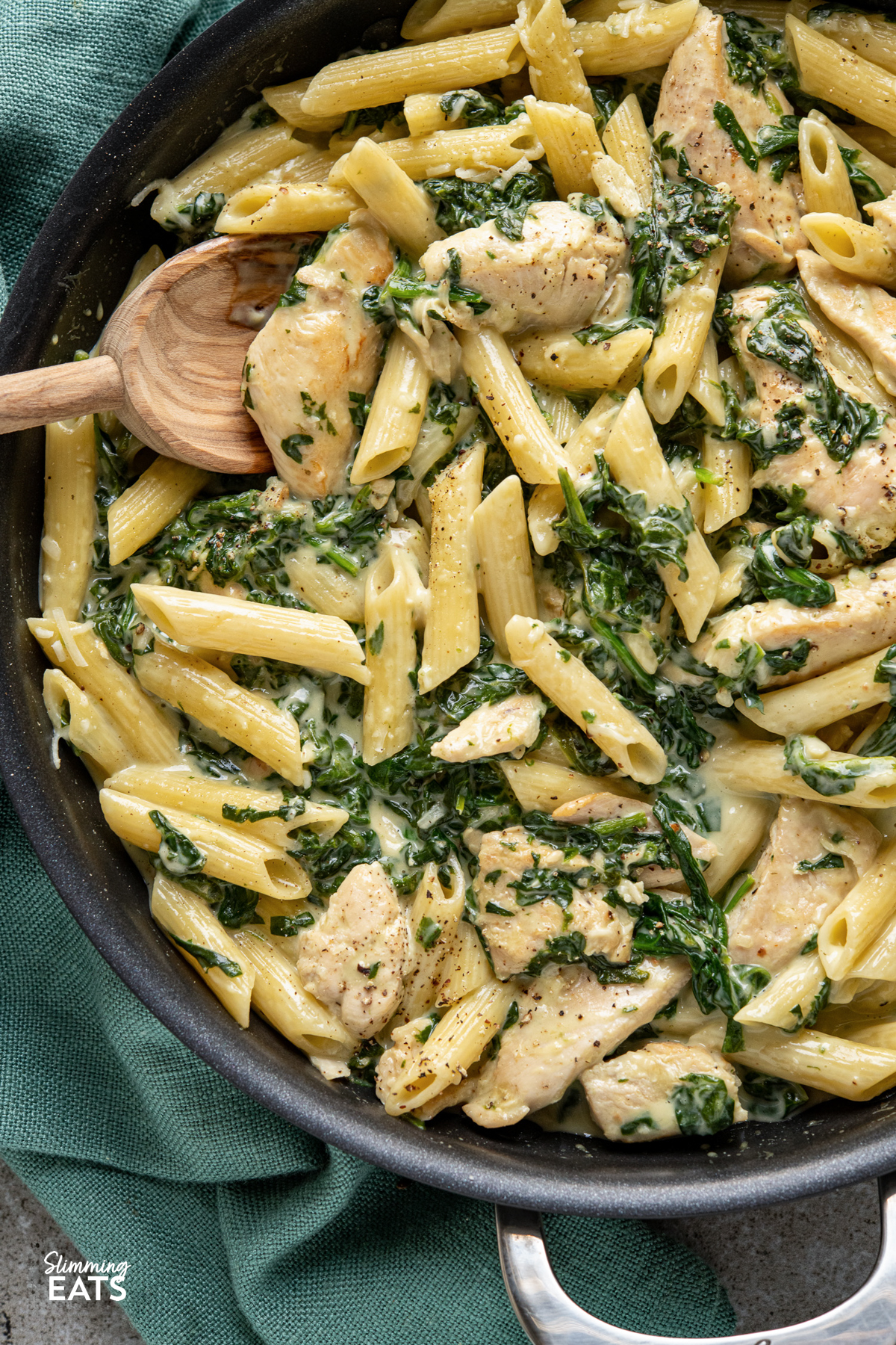 close up of creamy garlic chicken with spinach penne pasta in black frying pan with wooden spoon