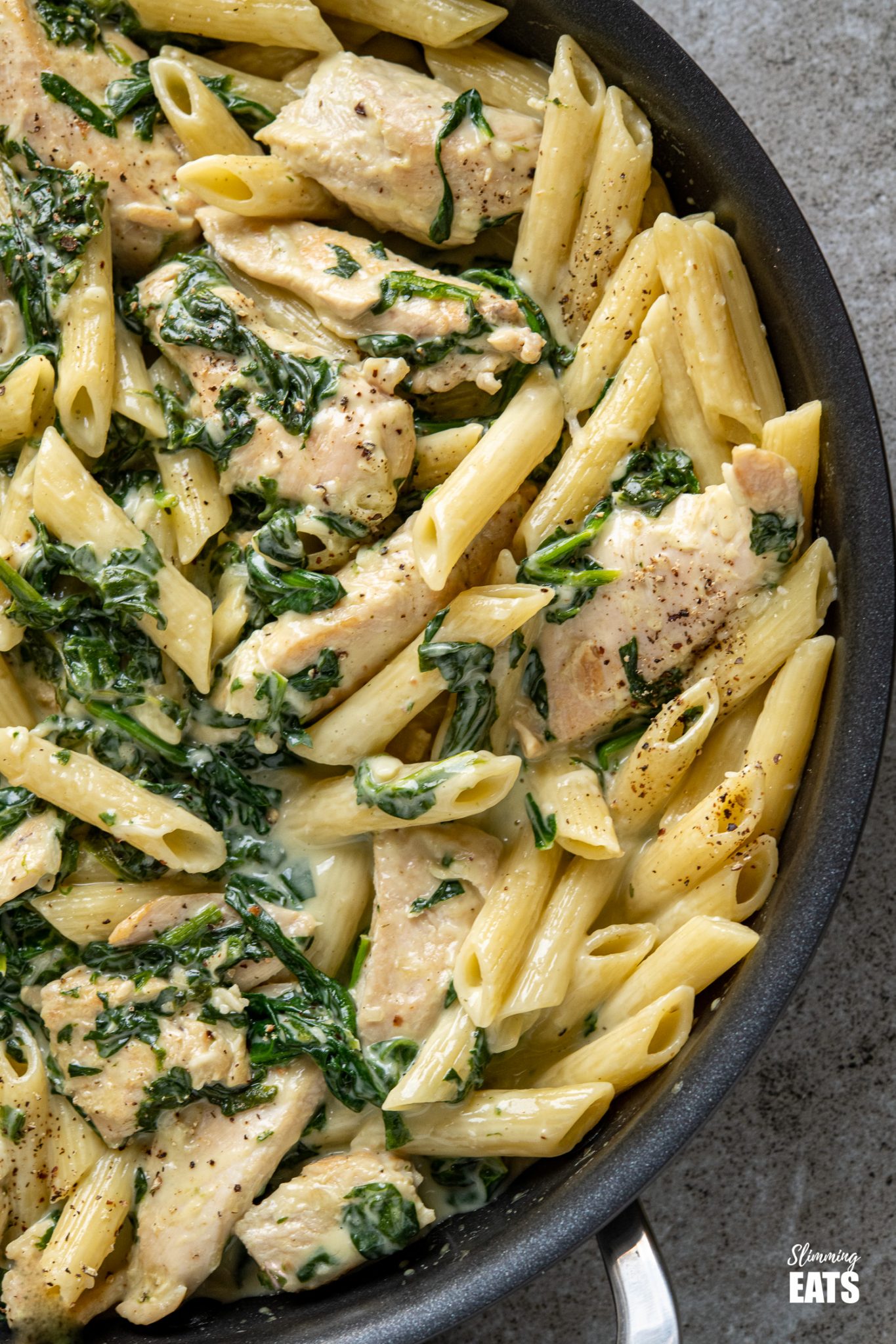 close up of creamy garlic chicken with spinach penne pasta in black frying pan with wooden spoon