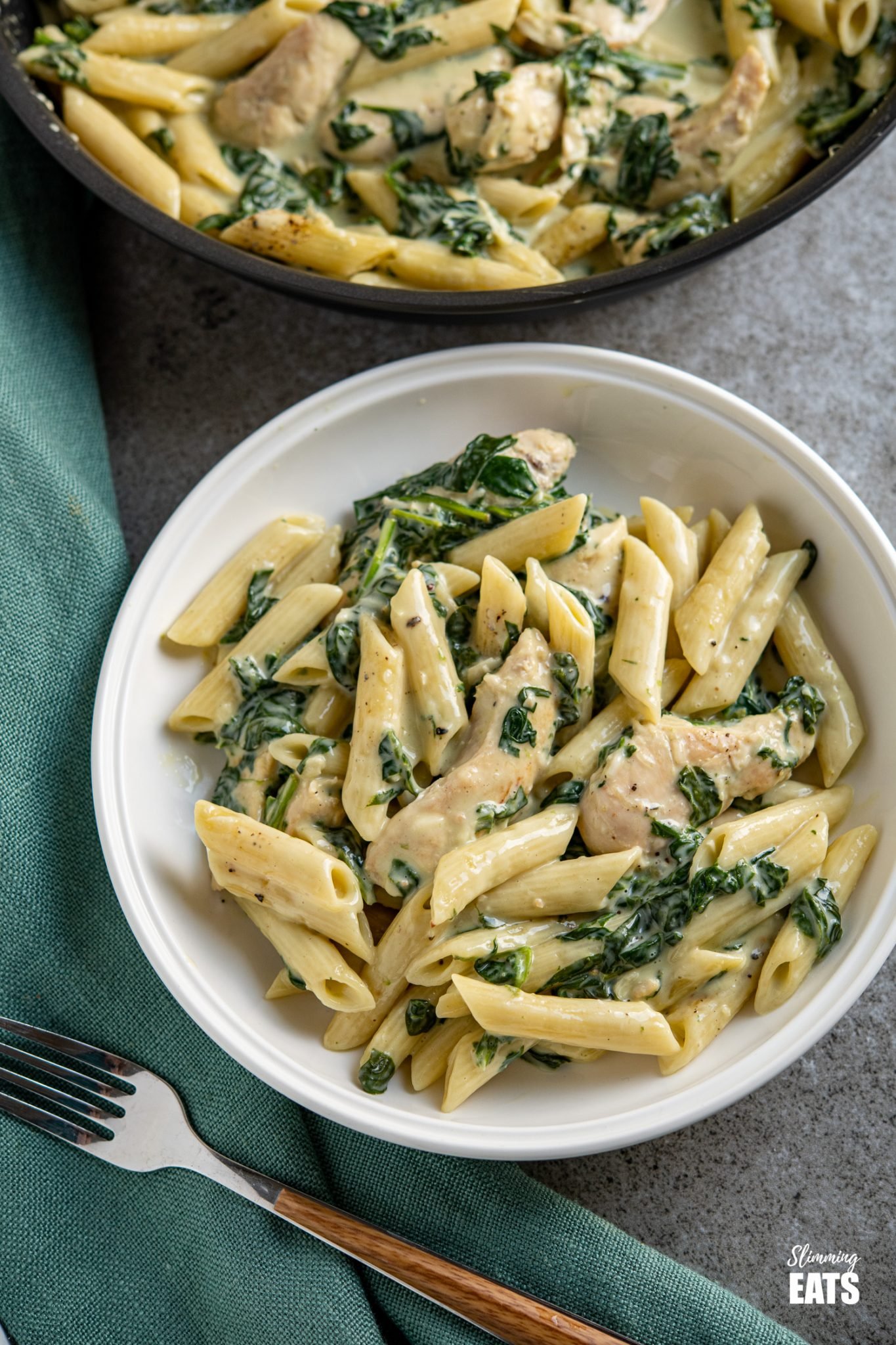 creamy garlic chicken with spinach penne pasta in white bowl with frying pan in background