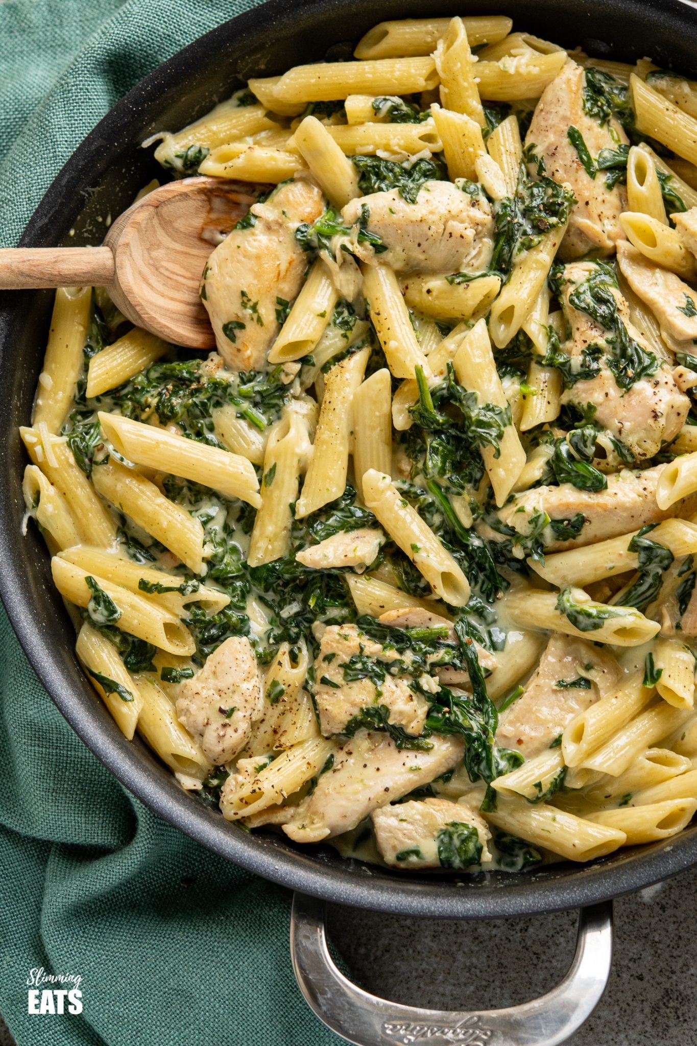 close up of creamy garlic chicken with spinach penne pasta in black frying pan with wooden spoon