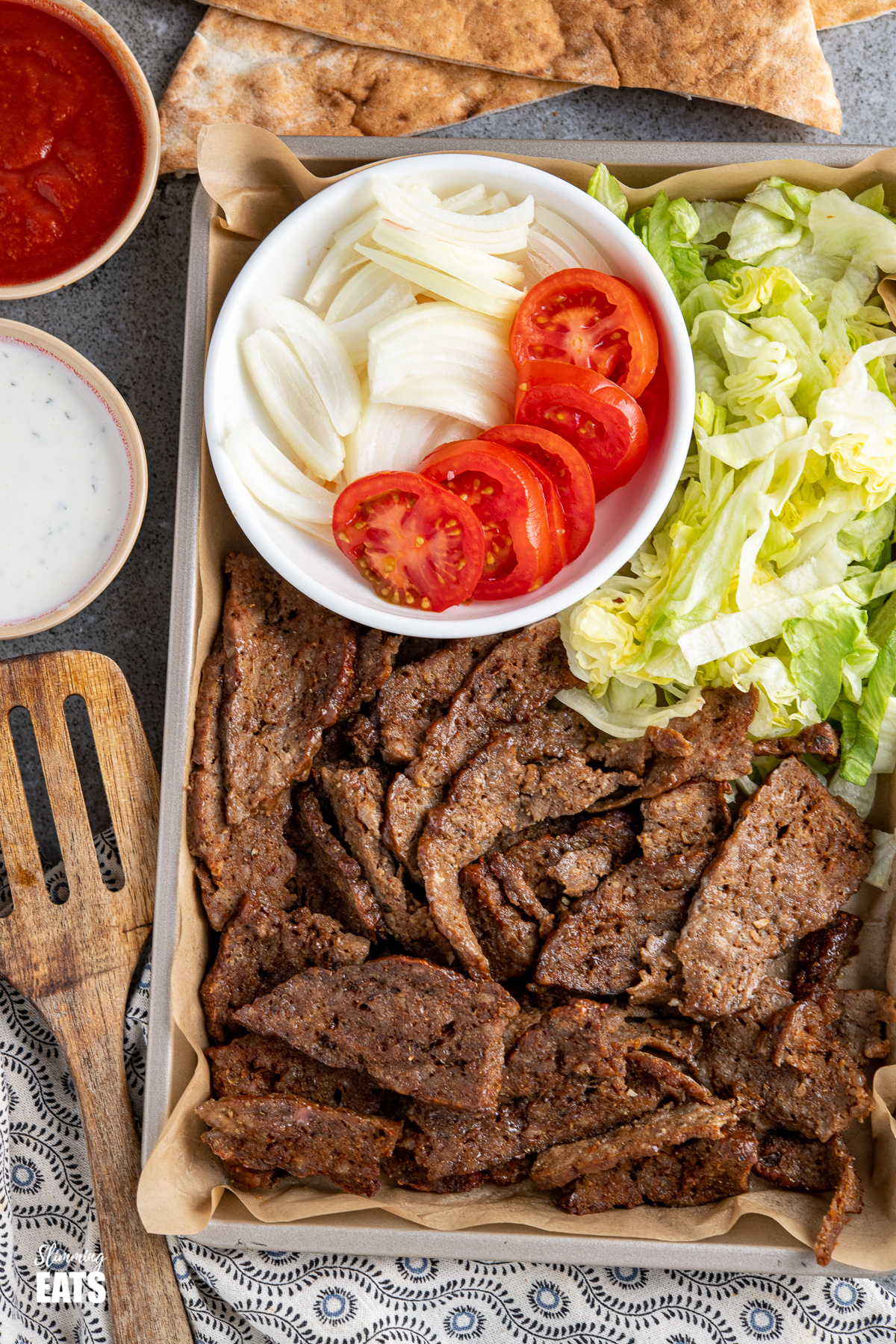 doner kebab meat on baking tray with salad and garlic sauce and wooden spatula to the left