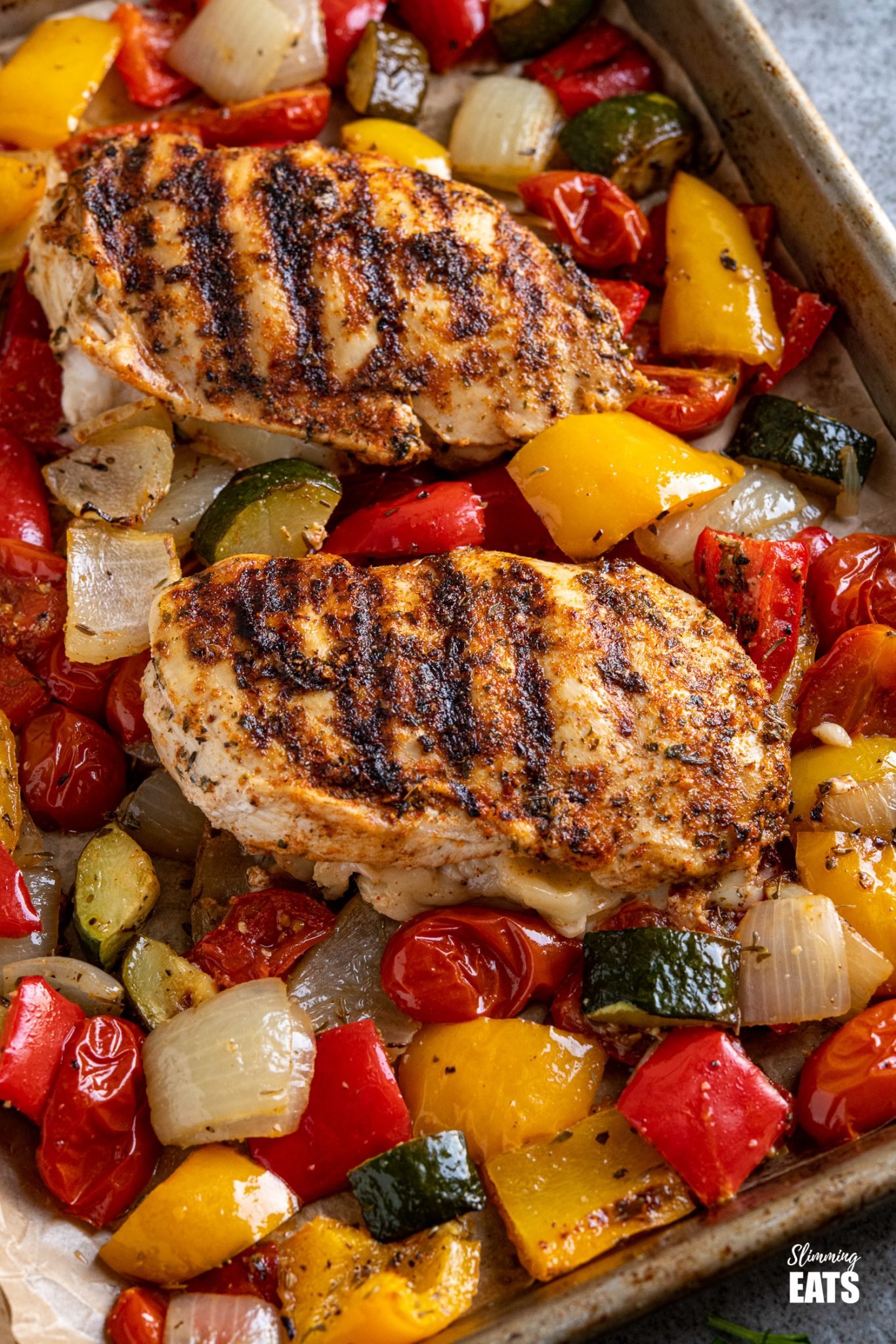 Simple seasoned Grilled Chicken Breast stuffed with mozzarella all delicious and melted served with a simple tray bake of roasted vegetables. #glutenfree #chicken #mozzarella #slimmingeats #weightwatchers #smartpoints #caloriecounting close up of two grilled chicken stuffed with mozzarella and roasted vegetables on a baking tray line with parchment paper