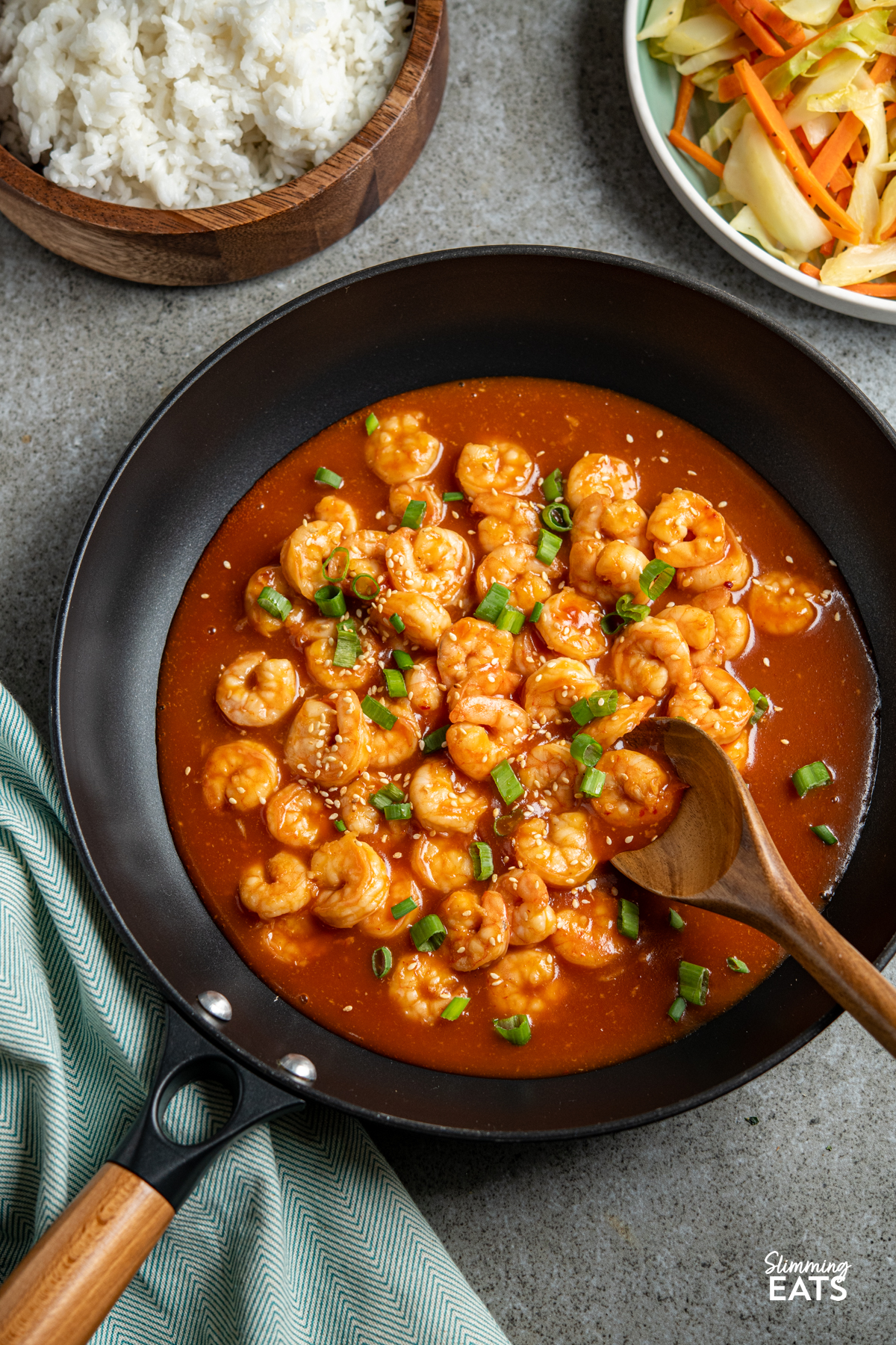 Sweet and Sour Shrimp - A super easy and quick recipe for shrimp in a Chinese style sweet and sour sauce, perfect with rice and some stir fried vegetables. #slimming #weightwatchers Sweet and sour shrimp in black frying pan with wooden spoon with wooden bowl with rice and white bbwl with veggies in background