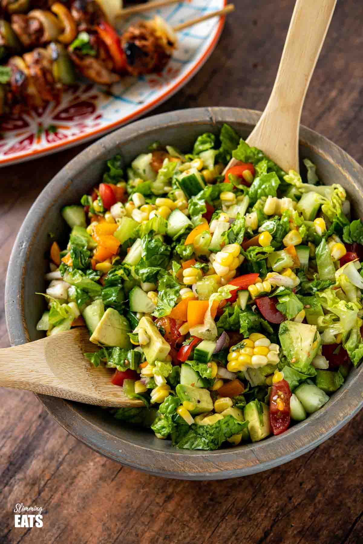 chopped salad in wooden bowl with wooden spoons with plate of chicken skewers behind