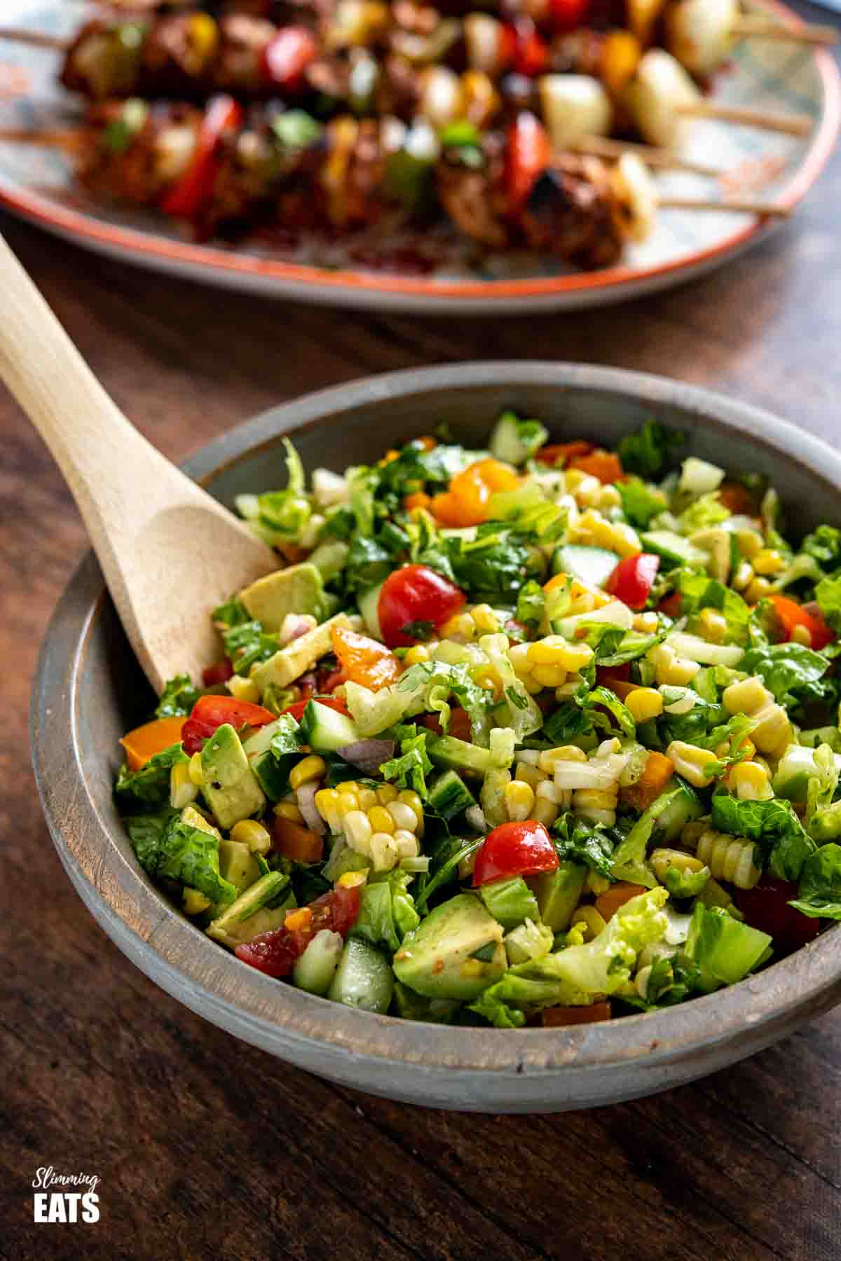 chopped salad in wooden bowl with wooden spoons with plate of chicken skewers behind