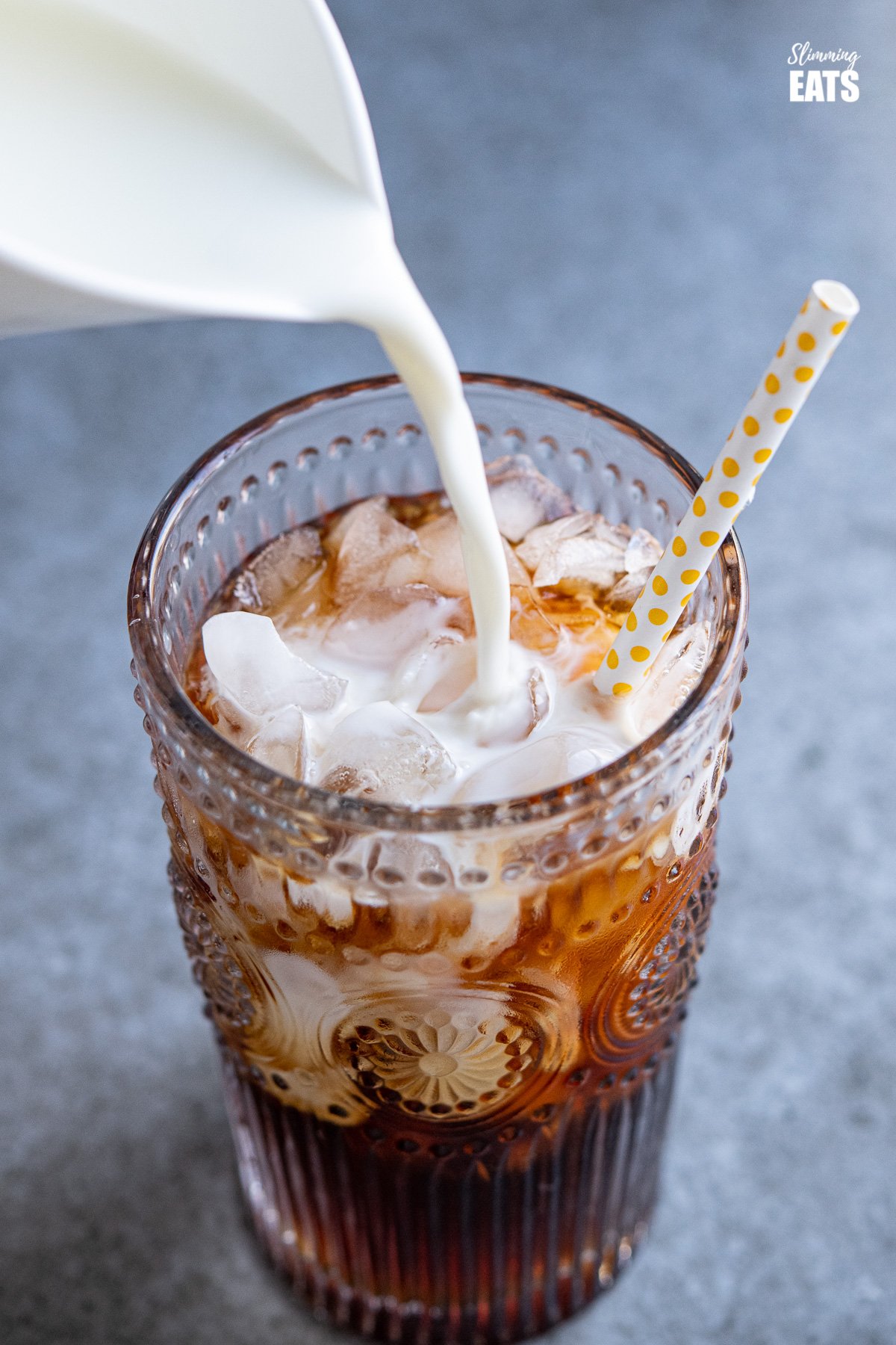 milk being poured into cold brew coffee with ice in a glass with yellow spotted white straw.
